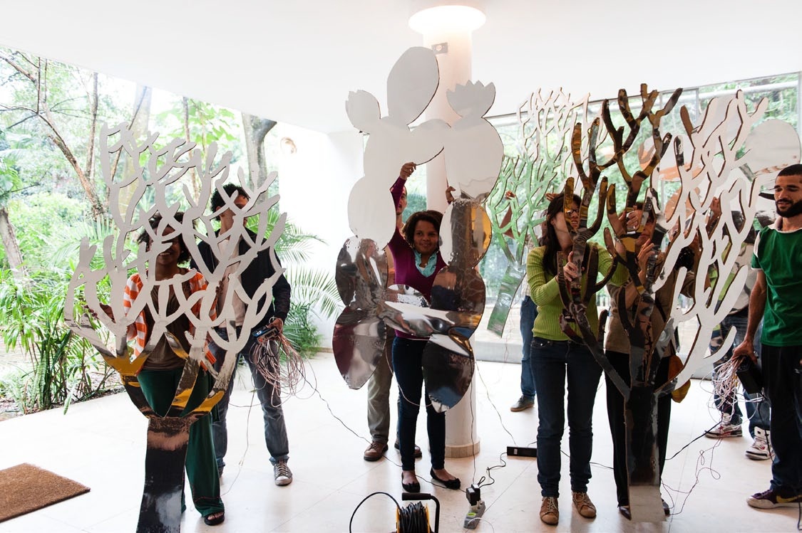 People in a bright, open gallery space with plants hold up mirrors shaped like cactuses.
