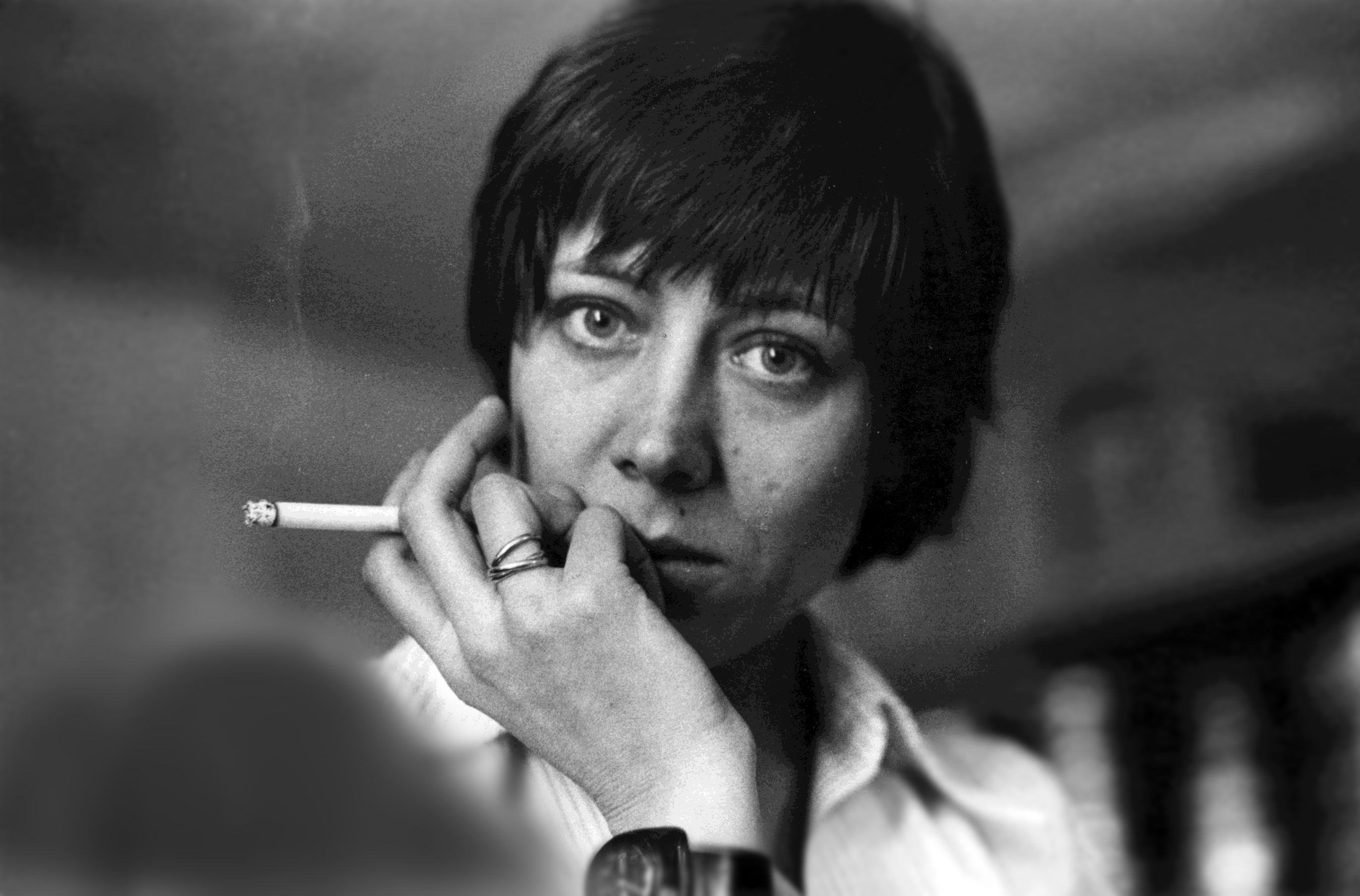 Close up of a woman with short dark hair, looking at the camera. Her fingers hold a lit cigarette and curl against her mouth.