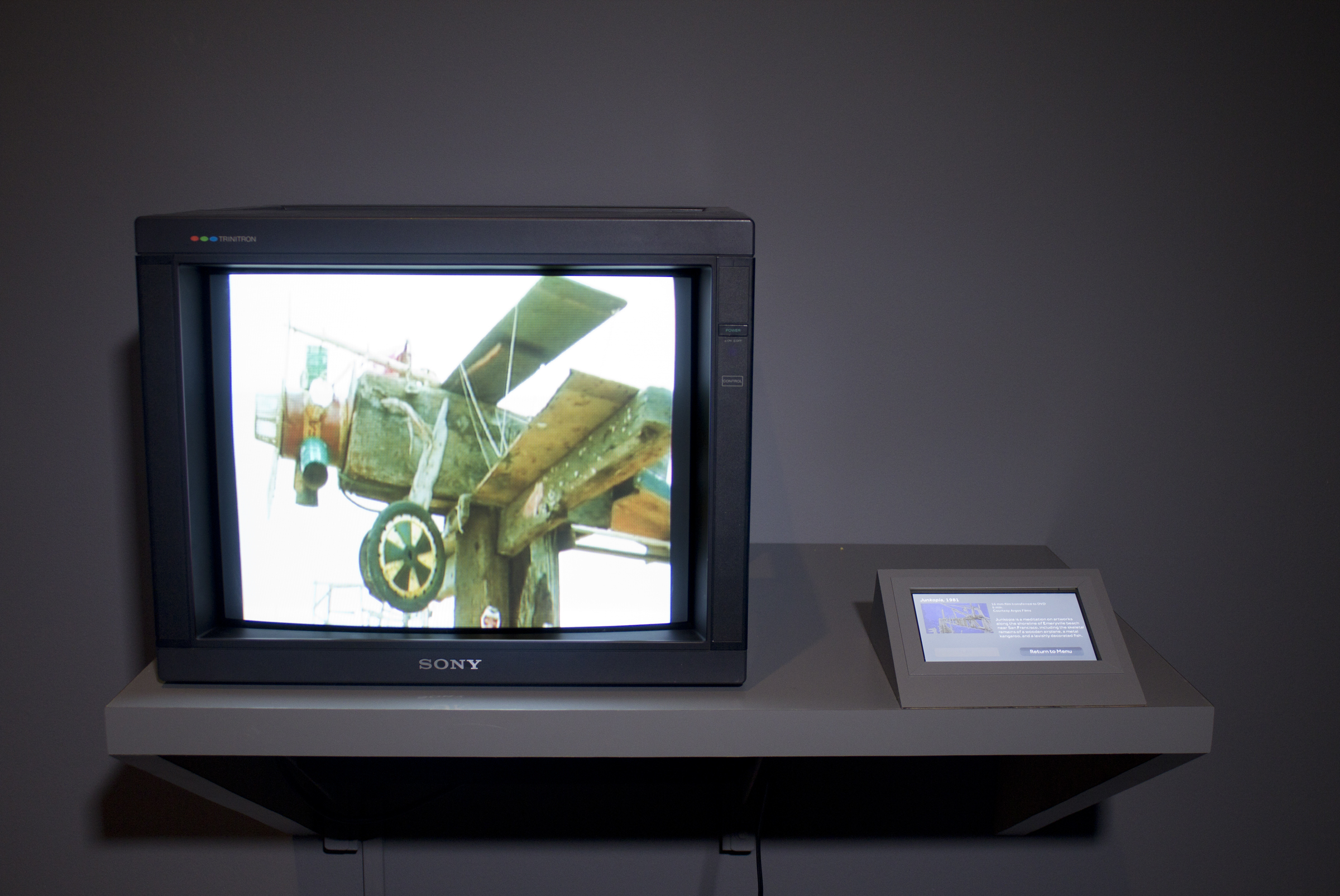 A monitor on a stand plays a video, black and white photos line the rear wall, and 5 photos hang on the wall at right.