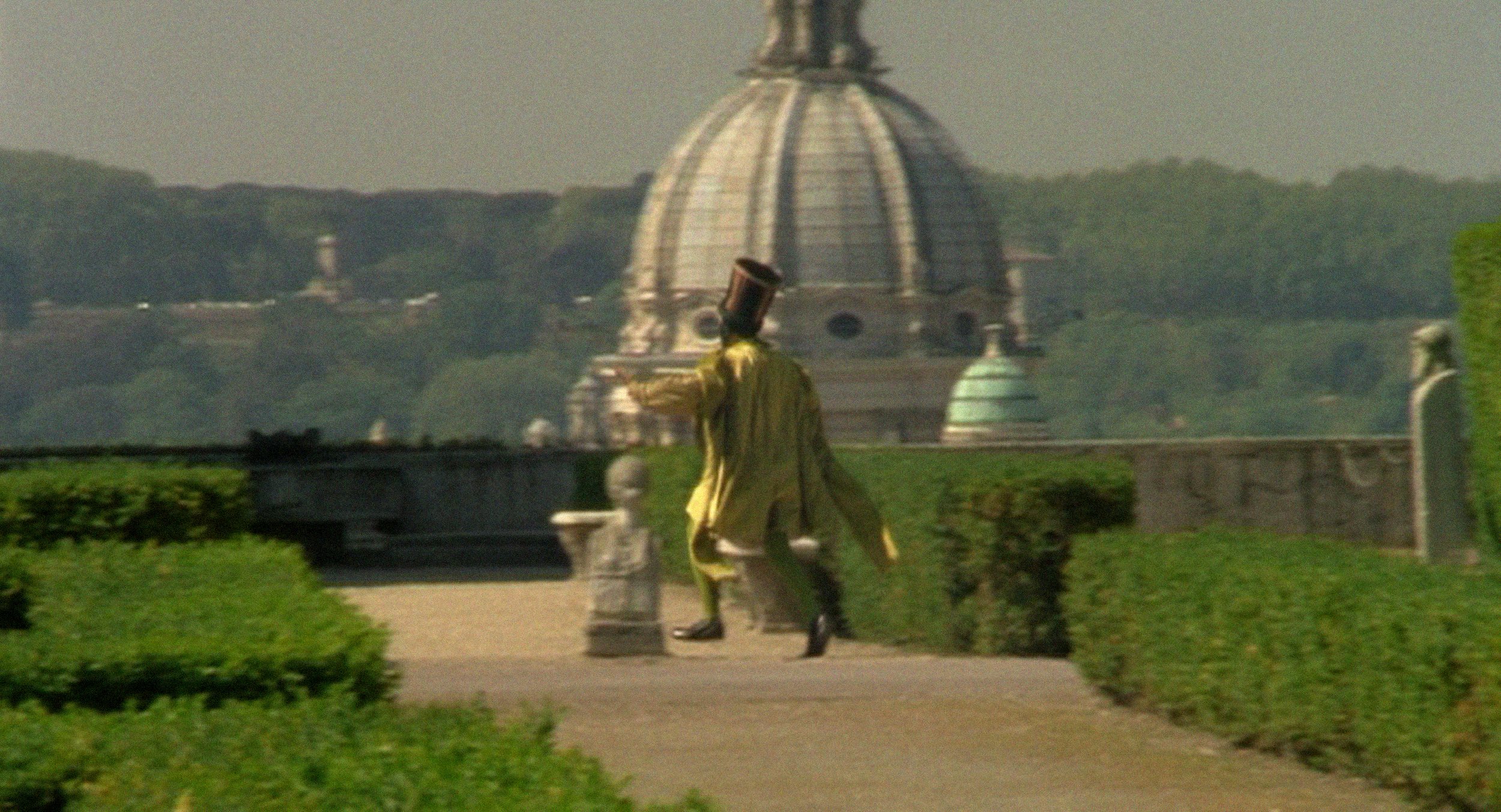 A man wearing a tunic and tall hat hurries away past hedgerows. An ornate white dome and mountains are seen in the background.