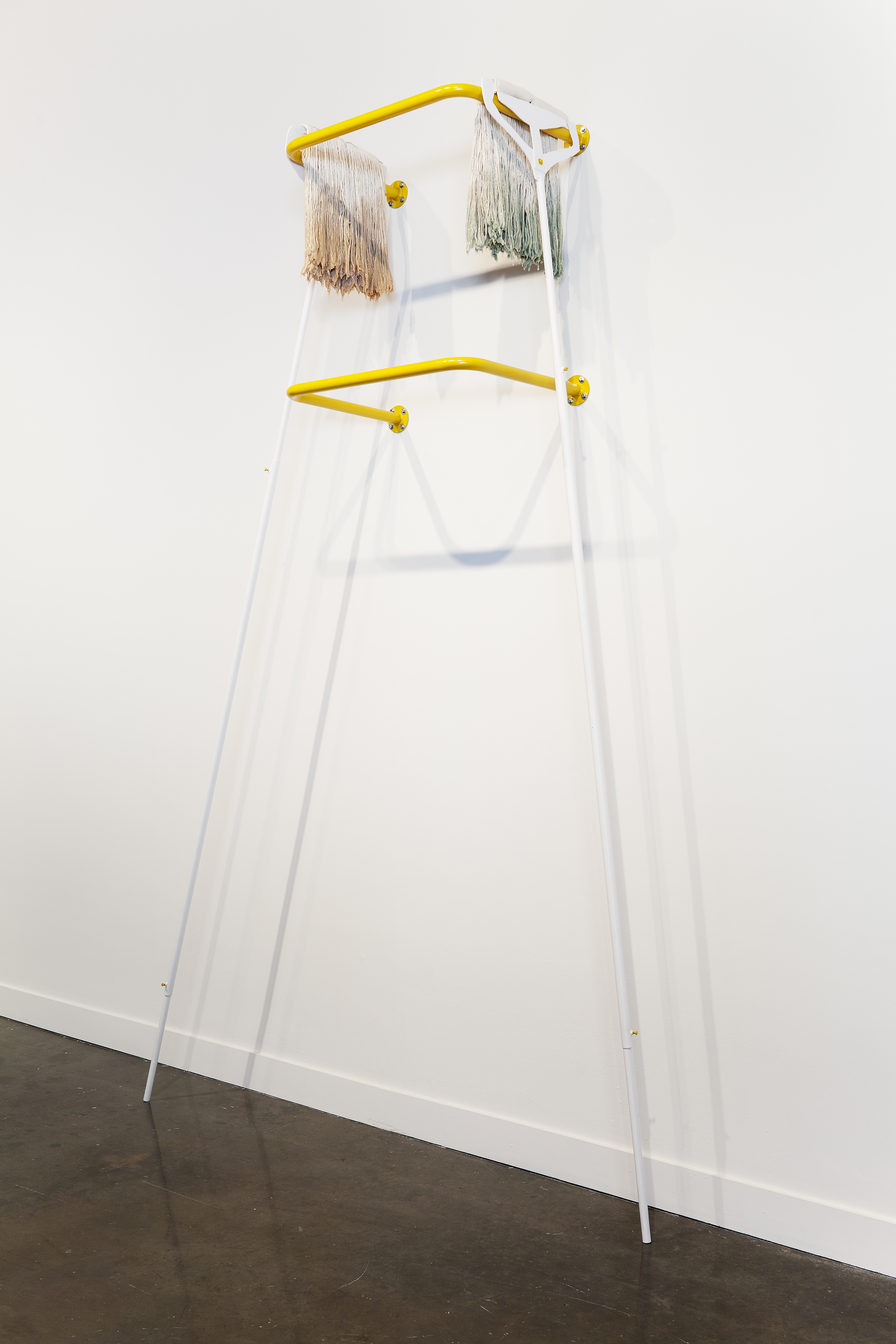 Two yellow railings mounted one over the other support 2 long white handled mops, heads draped over the sides of the top bar.