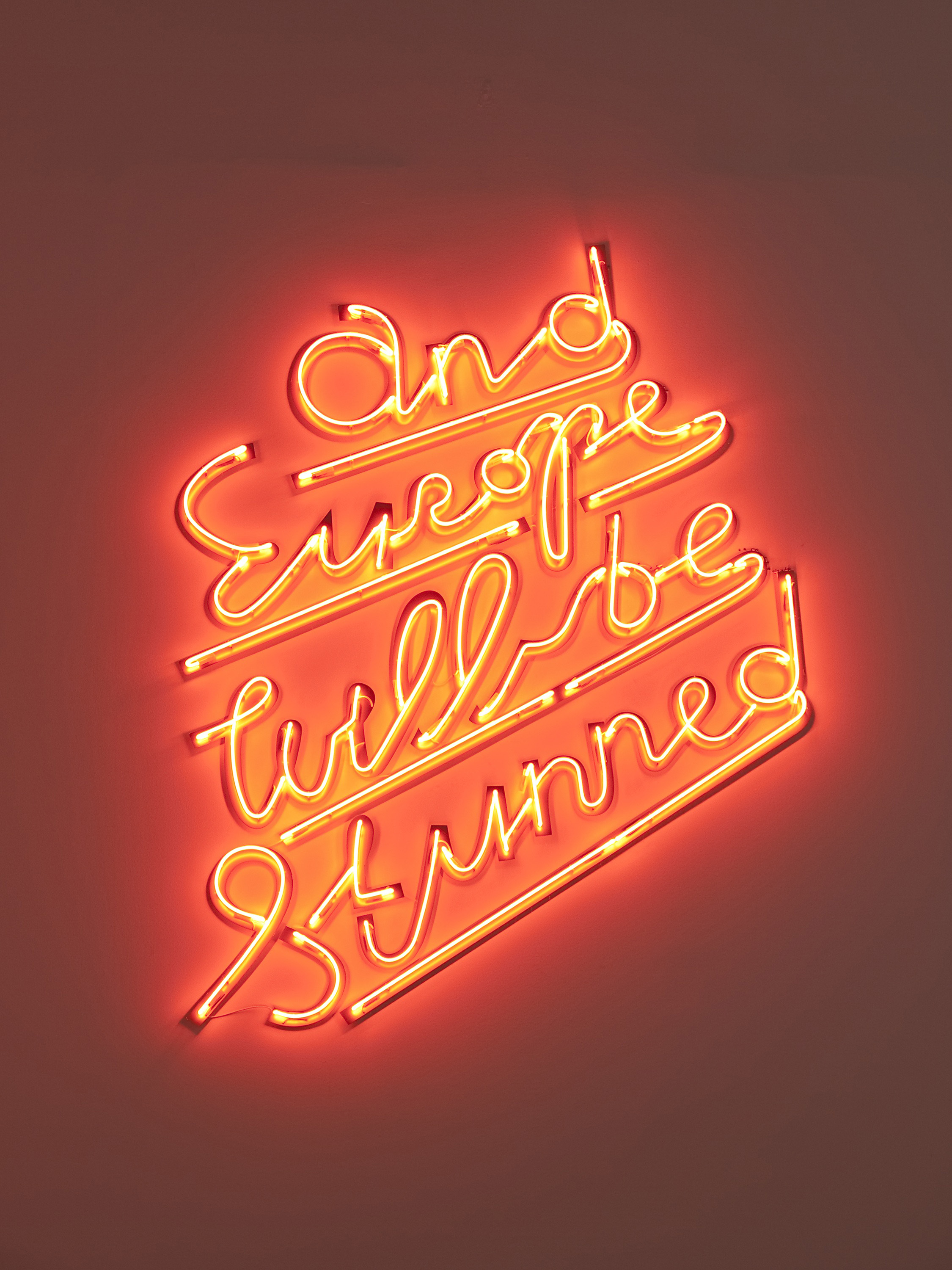 The words, “and Europe will be Stunned” in neon, glows orange red on a wall.