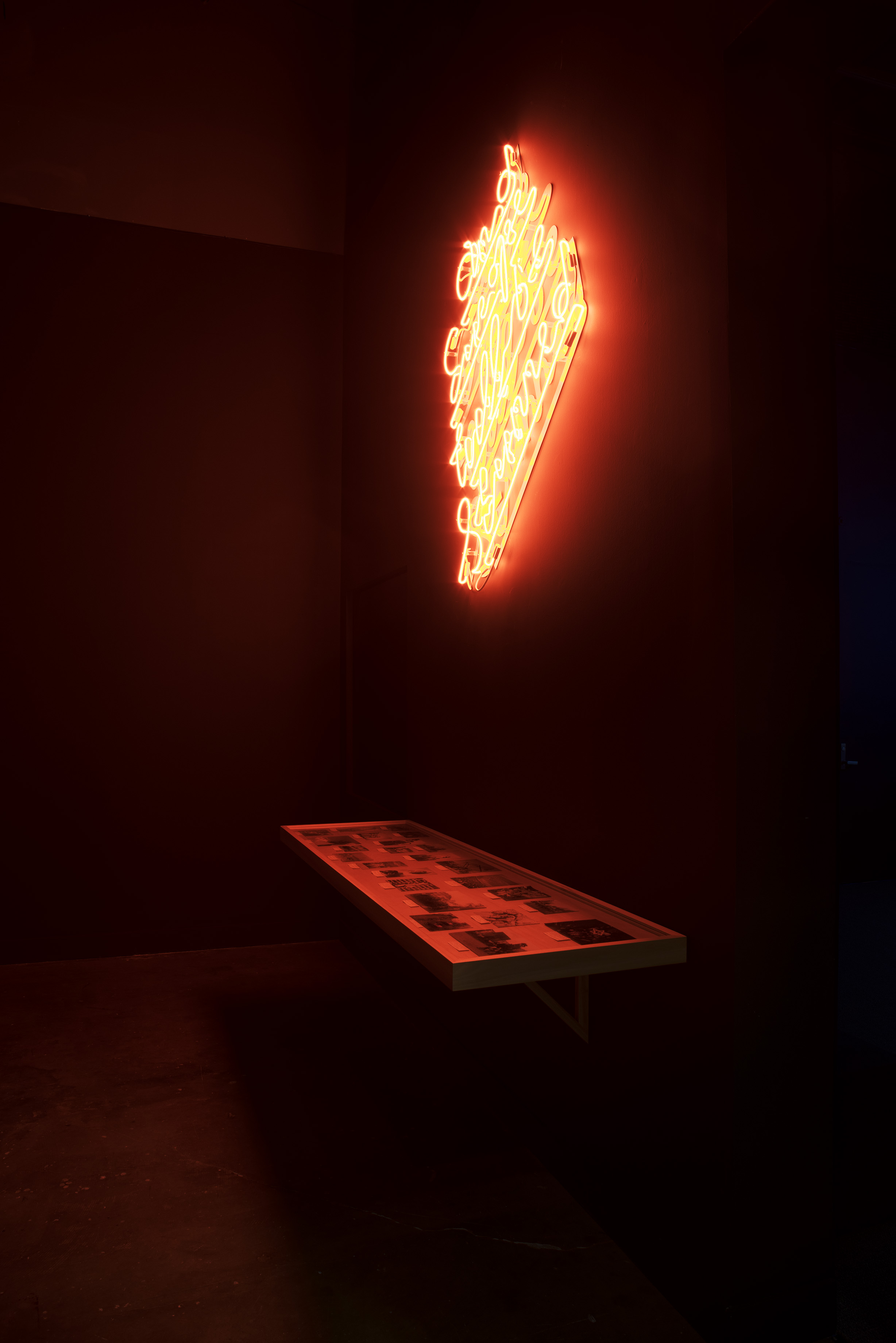 Neon art on the wall spill red light onto vitrine holding documents and photographs below in a dark gallery.
