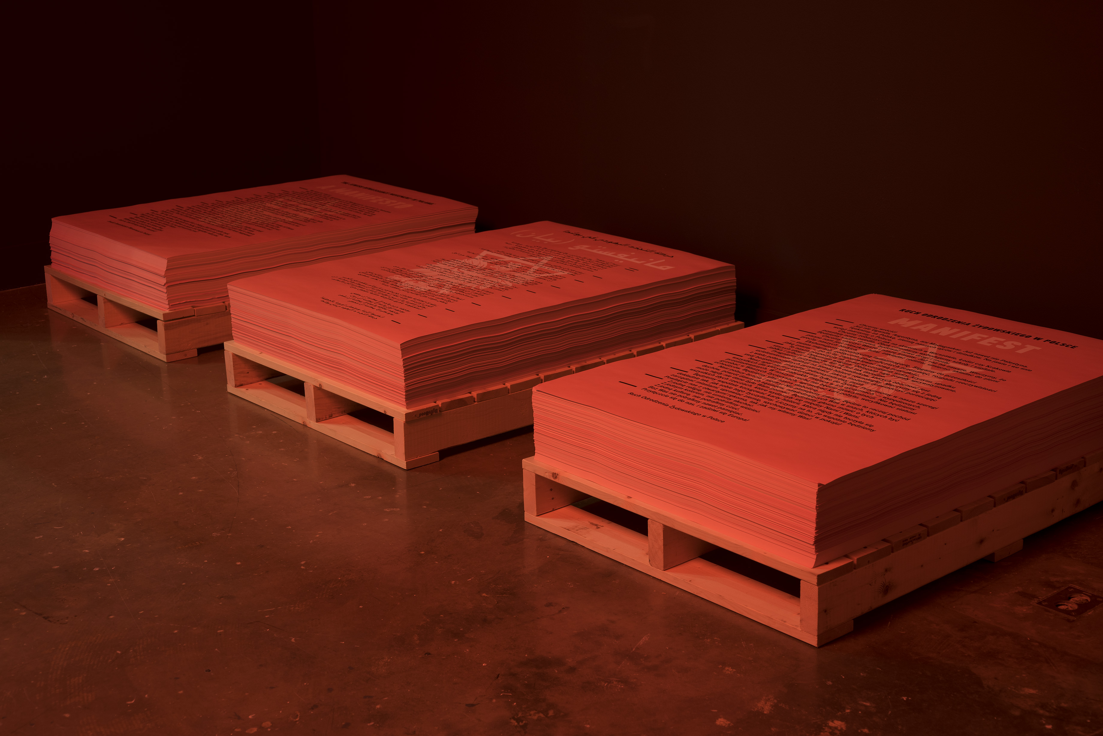 Bathed in red light in a dark gallery, 3 framed documents rest on 3 pallets on the gallery floor.