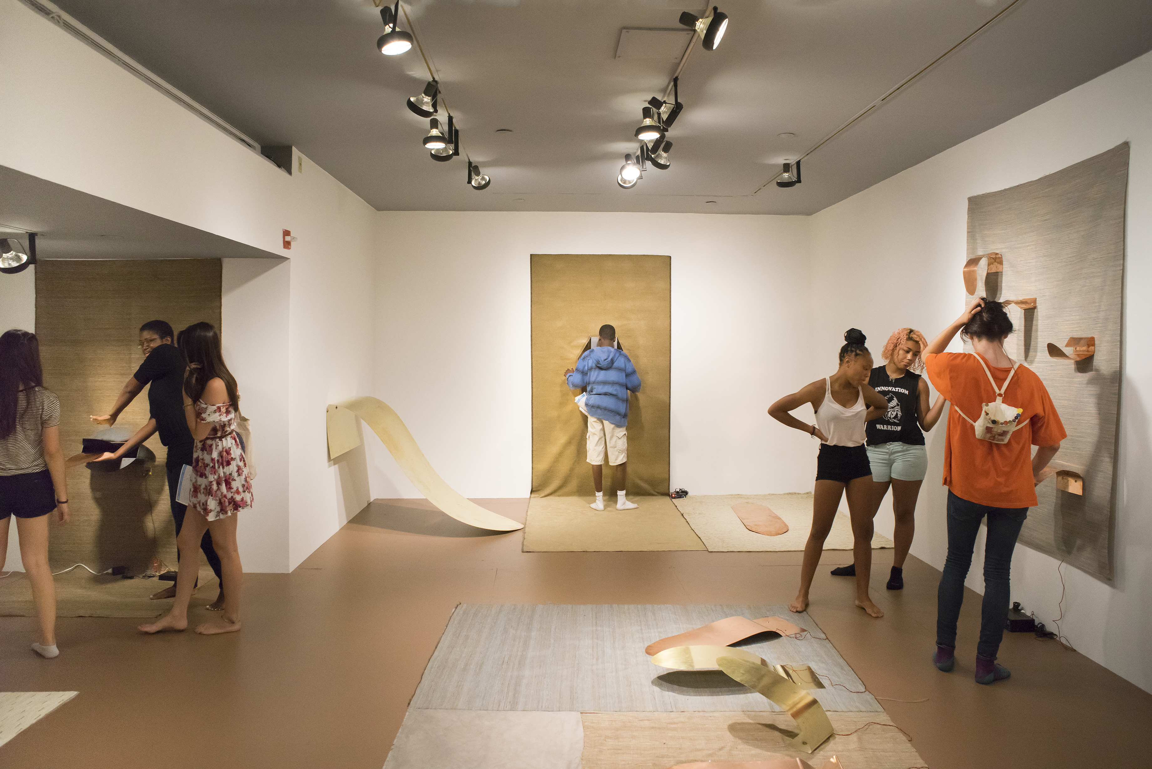 Many young visitors in bare feet interact with metal sculptures mounted on textured fabrics on the floor and the wall.