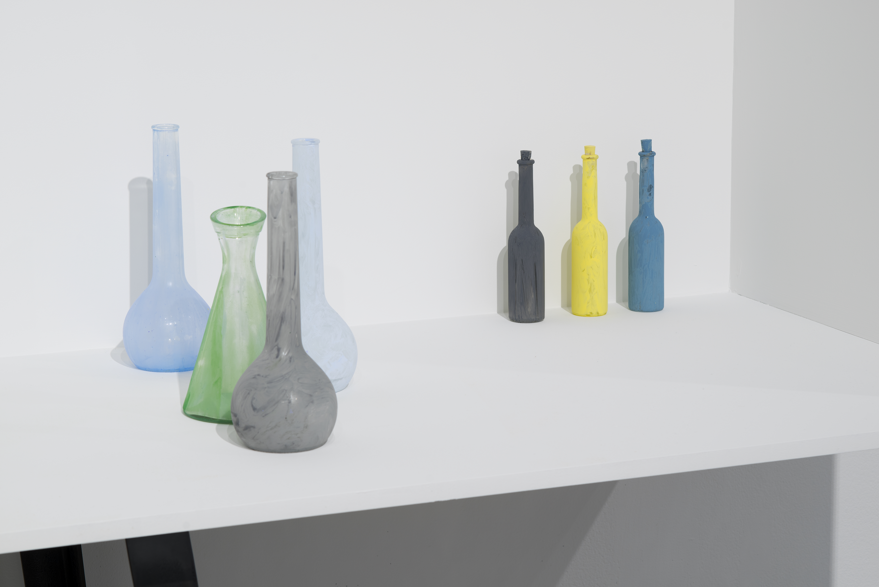 Two groupings of painted bottles on a white shelf. One grouping, to the right are stopped, while the other grouping is open.