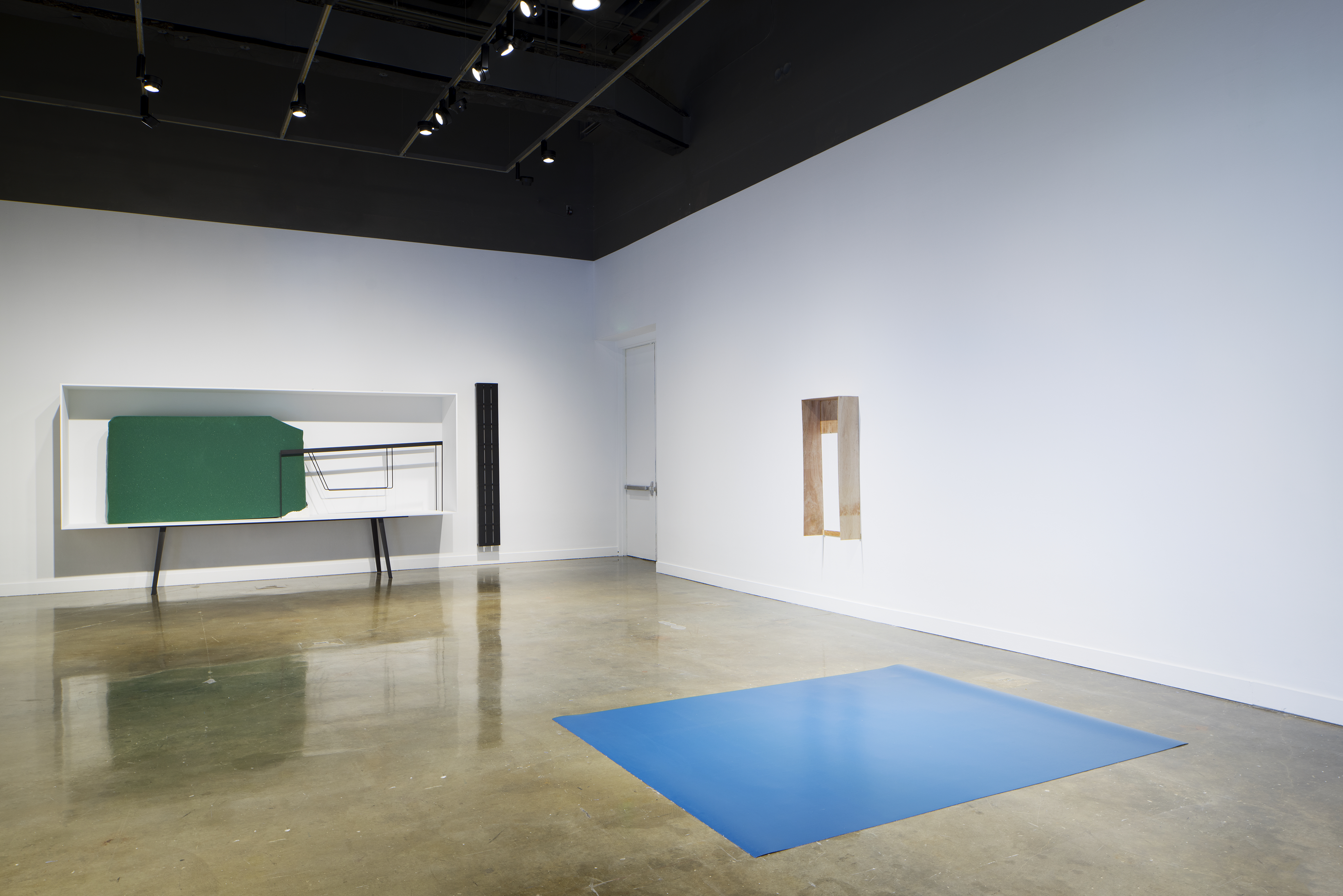 A bright blue mat on the ground, a wooden object on the right wall, and a green and metal sculpture in a white display case.