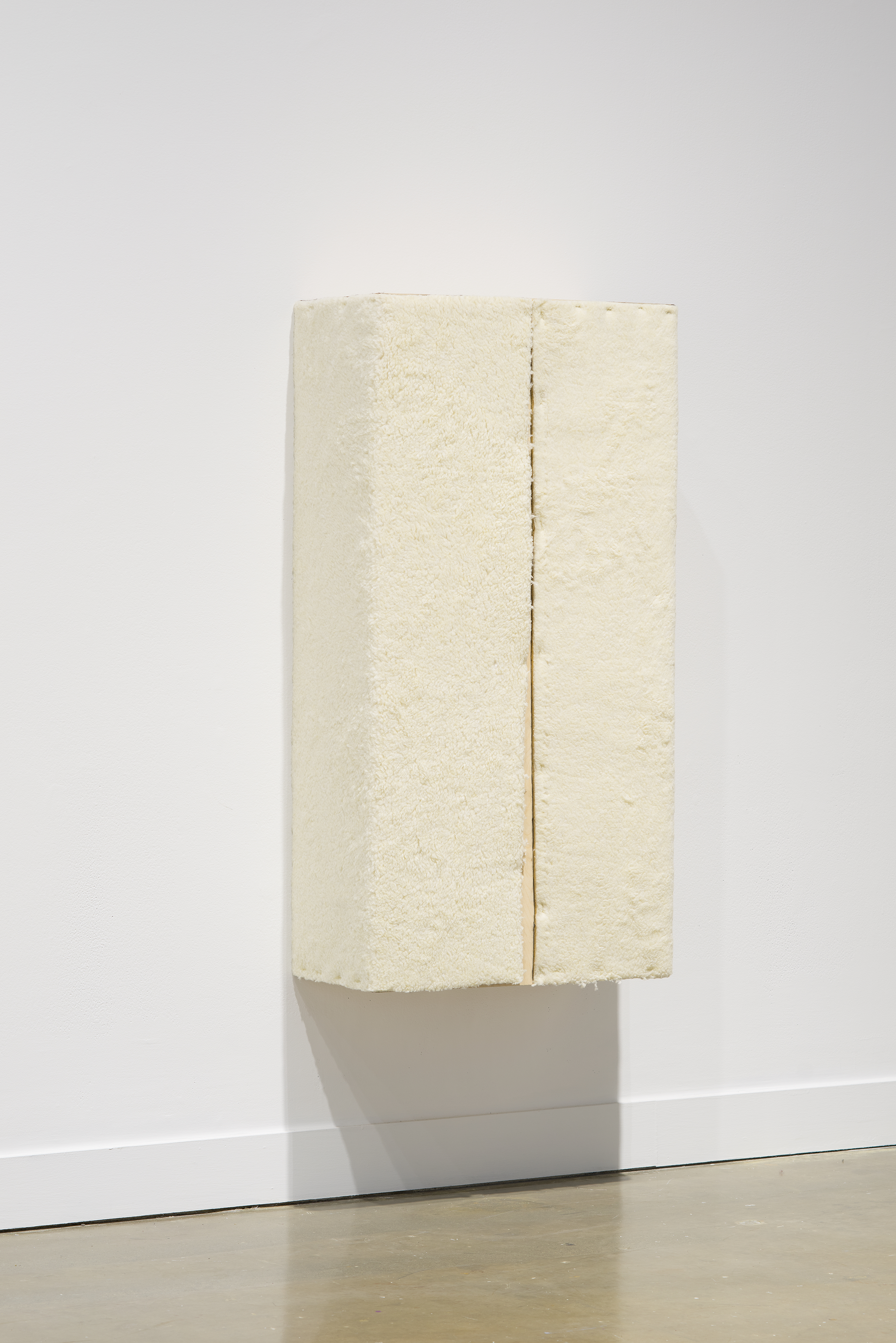 A wall-mounted, cream colored sculpture with an orange line down the middle casts a shadow on the wall and floor.