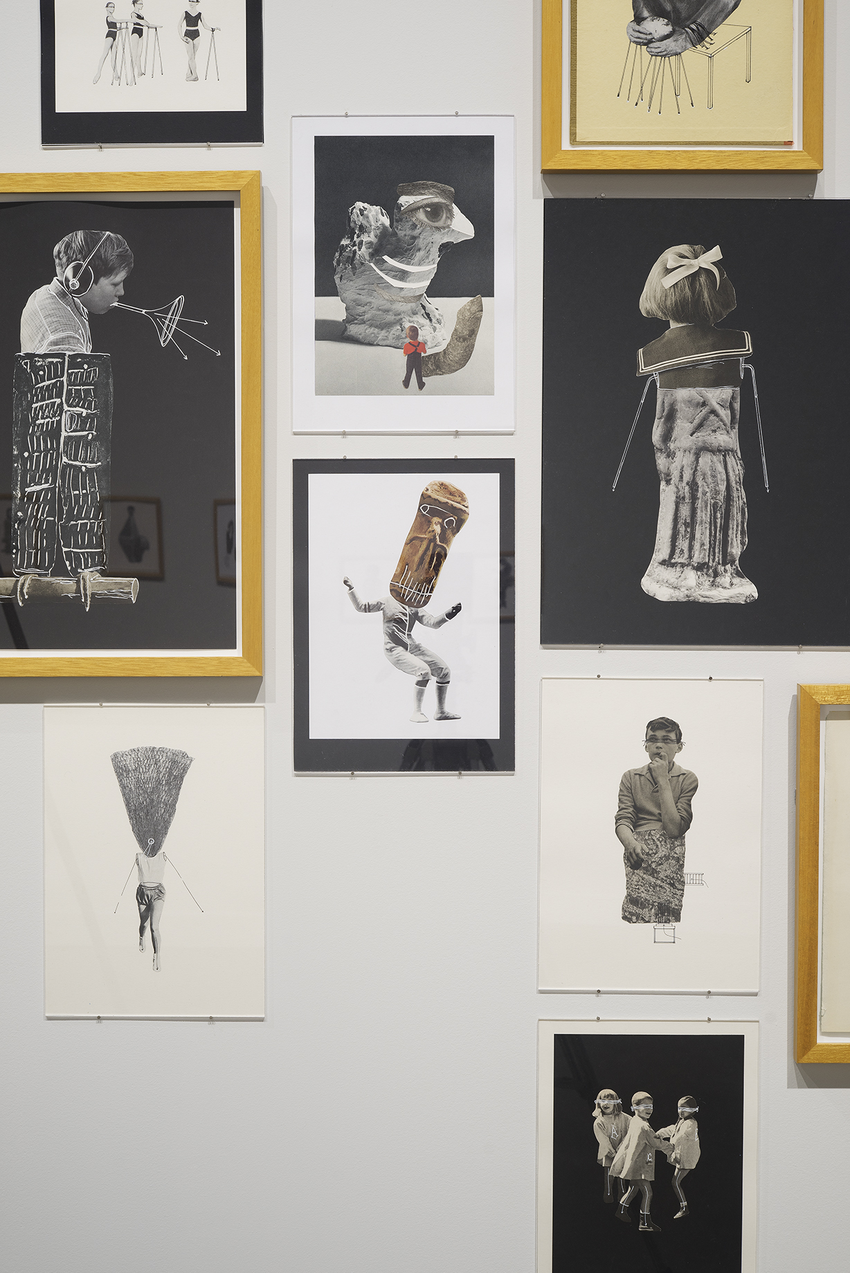 A number of collages of people hang on a wall, some framed and matted, others only matted and rest simply on paper.