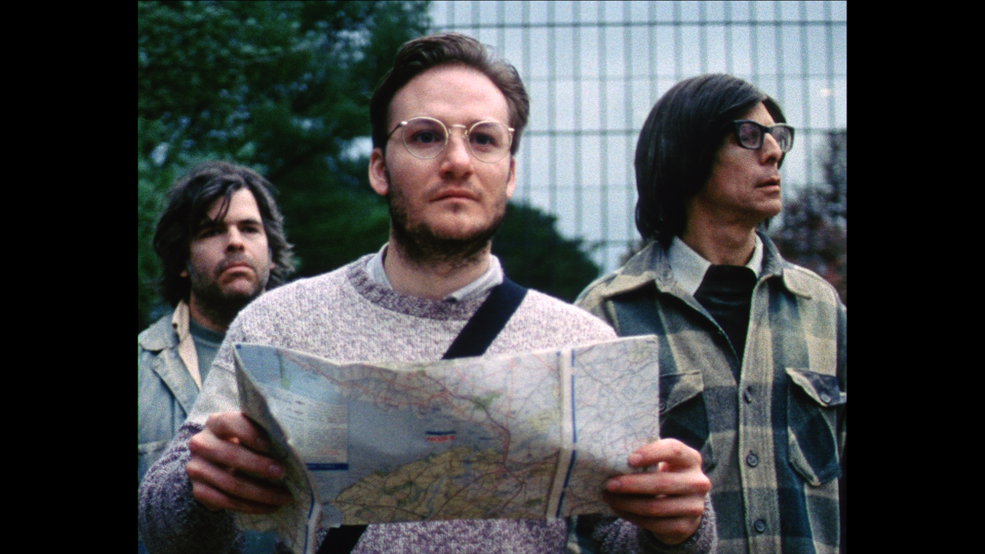 Three men, one in front of the other two, stand outside a mirrored building with landscaping. The man in front holds a map.