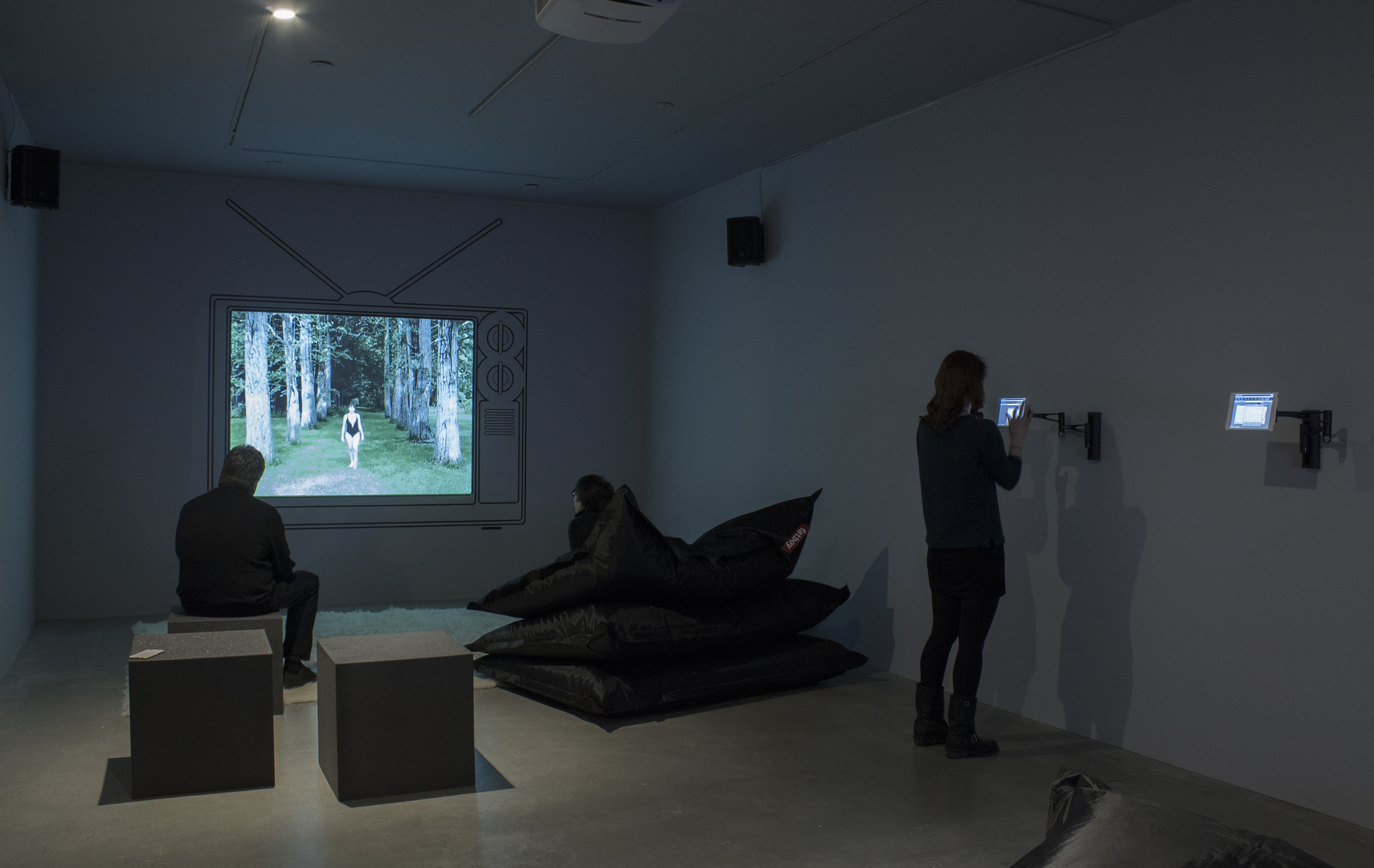 2 visitors watch a projected video framed by the drawing of an old TV, as another visitor interacts with a wall-mounted iPad.