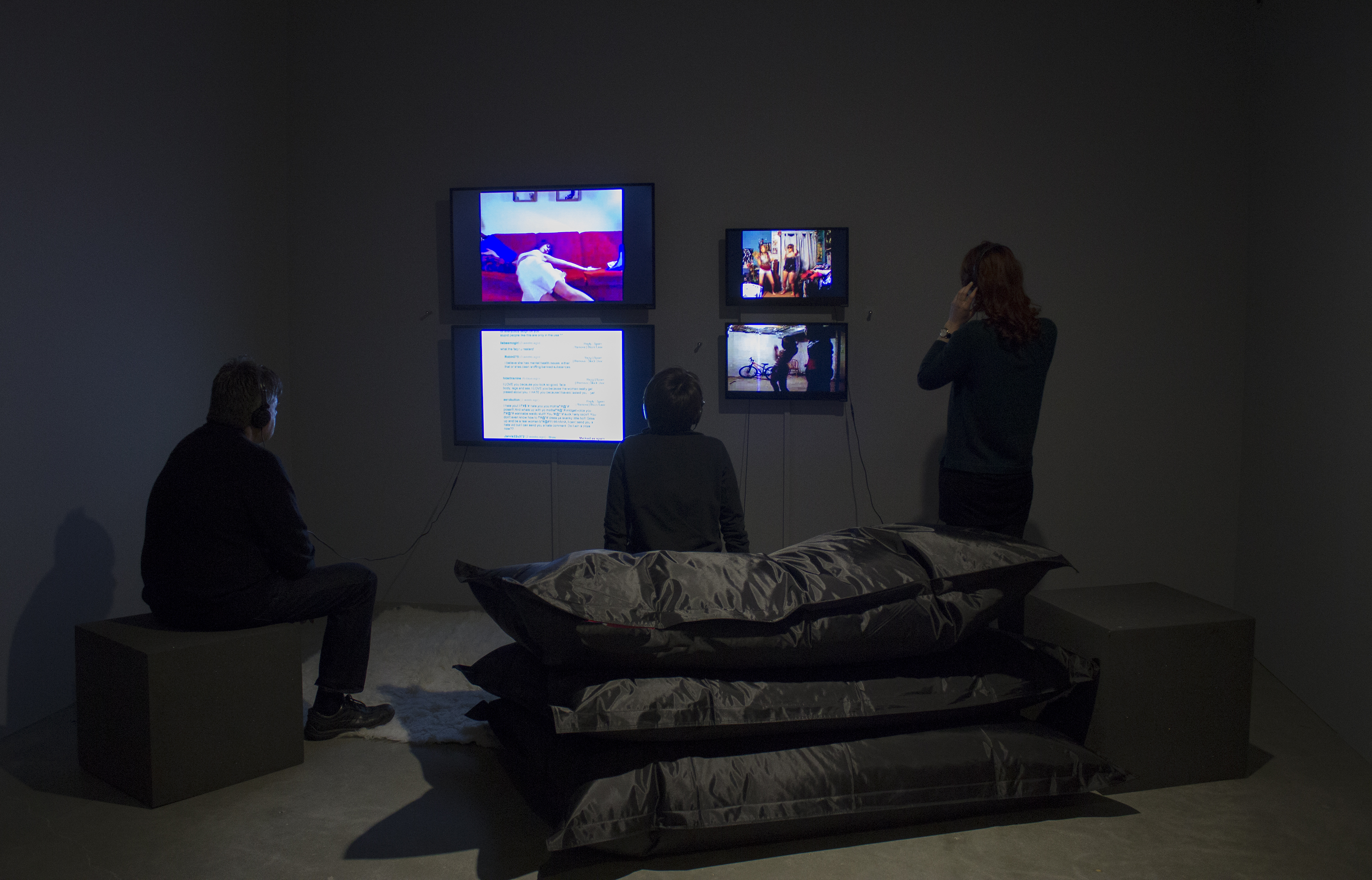 3 visitors, 1 standing, 2 seated on beanbags and stool, watch 4 wall-mounted monitors in a darkened gallery.