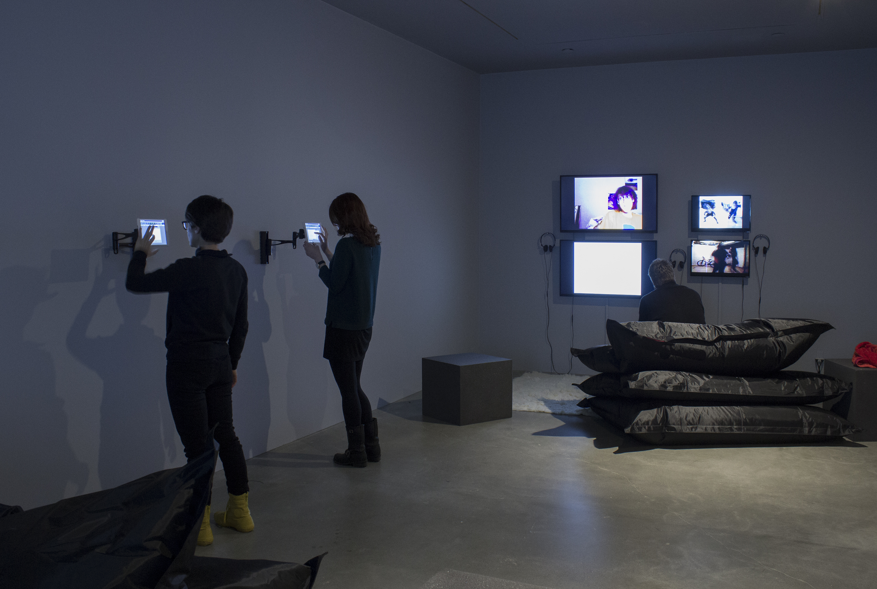 2 visitors interact with wall-mounted iPads while a visitor sits on beanbags watching 4 wall-mounted monitors.