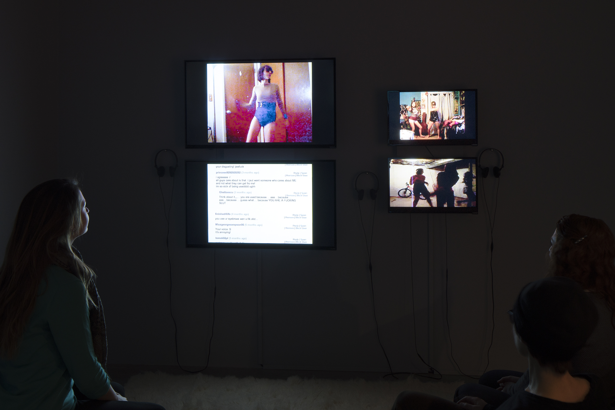 2 visitors watching videos on 4 wall-mounted monitors in a dark gallery.