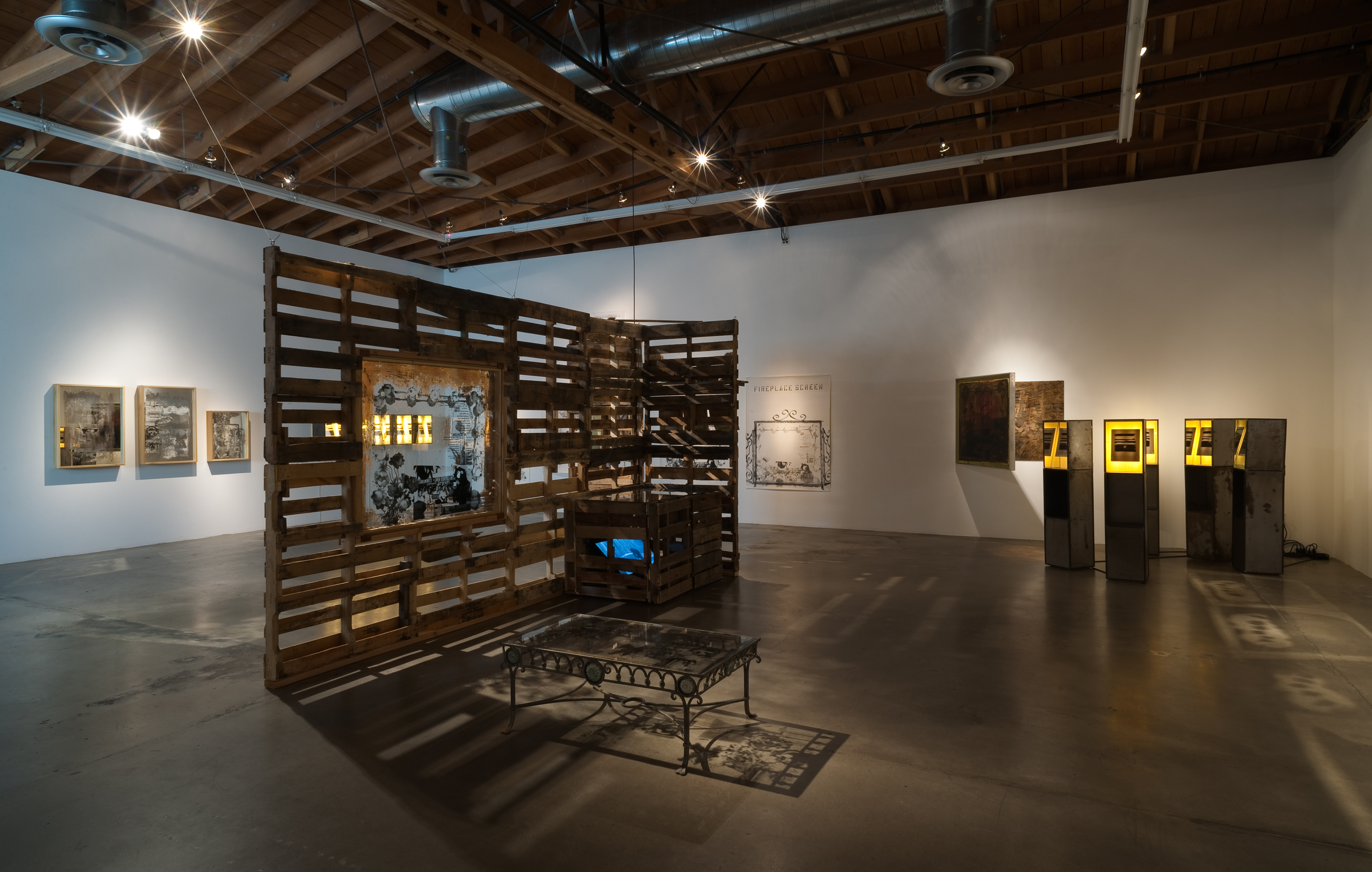 A wooden structure is situated in a gallery space with other artworks lit on the walls.