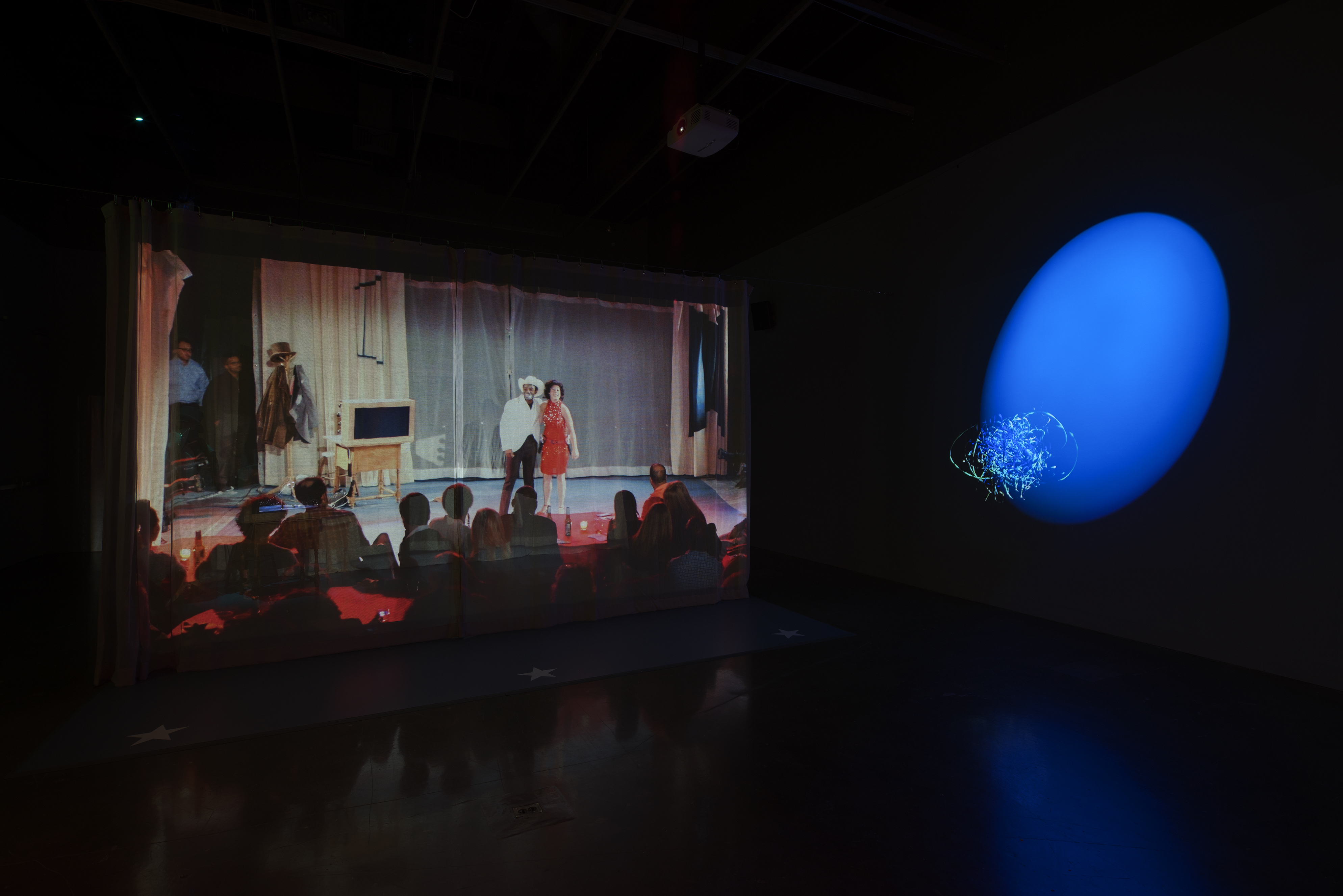 A dark space with a blue spotlight, a large projection of a woman and man performing with an audience in silhouette in front