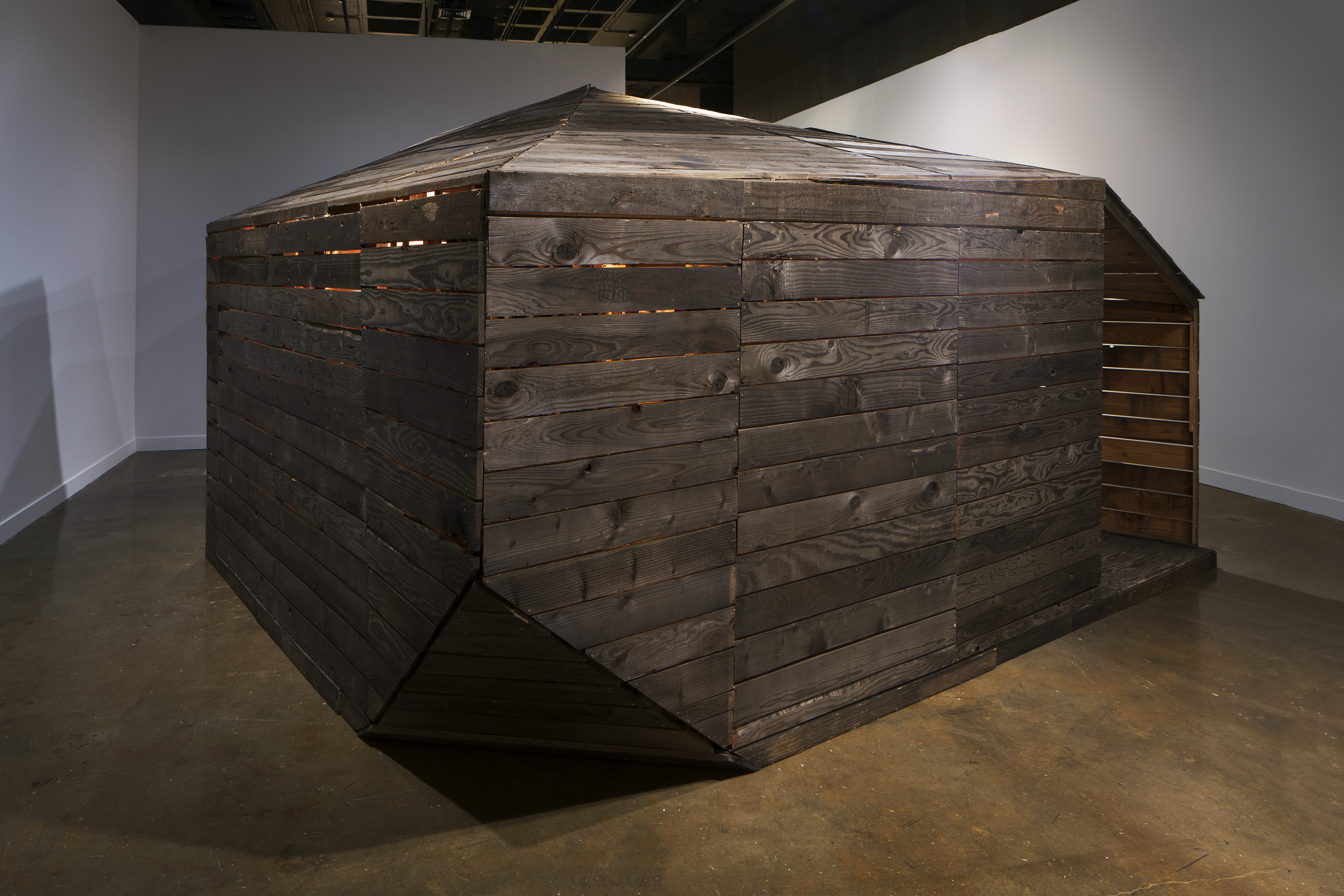 A large dark slatted wood cabin structure with pointed roof and lit from within, almost filling the space it’s in