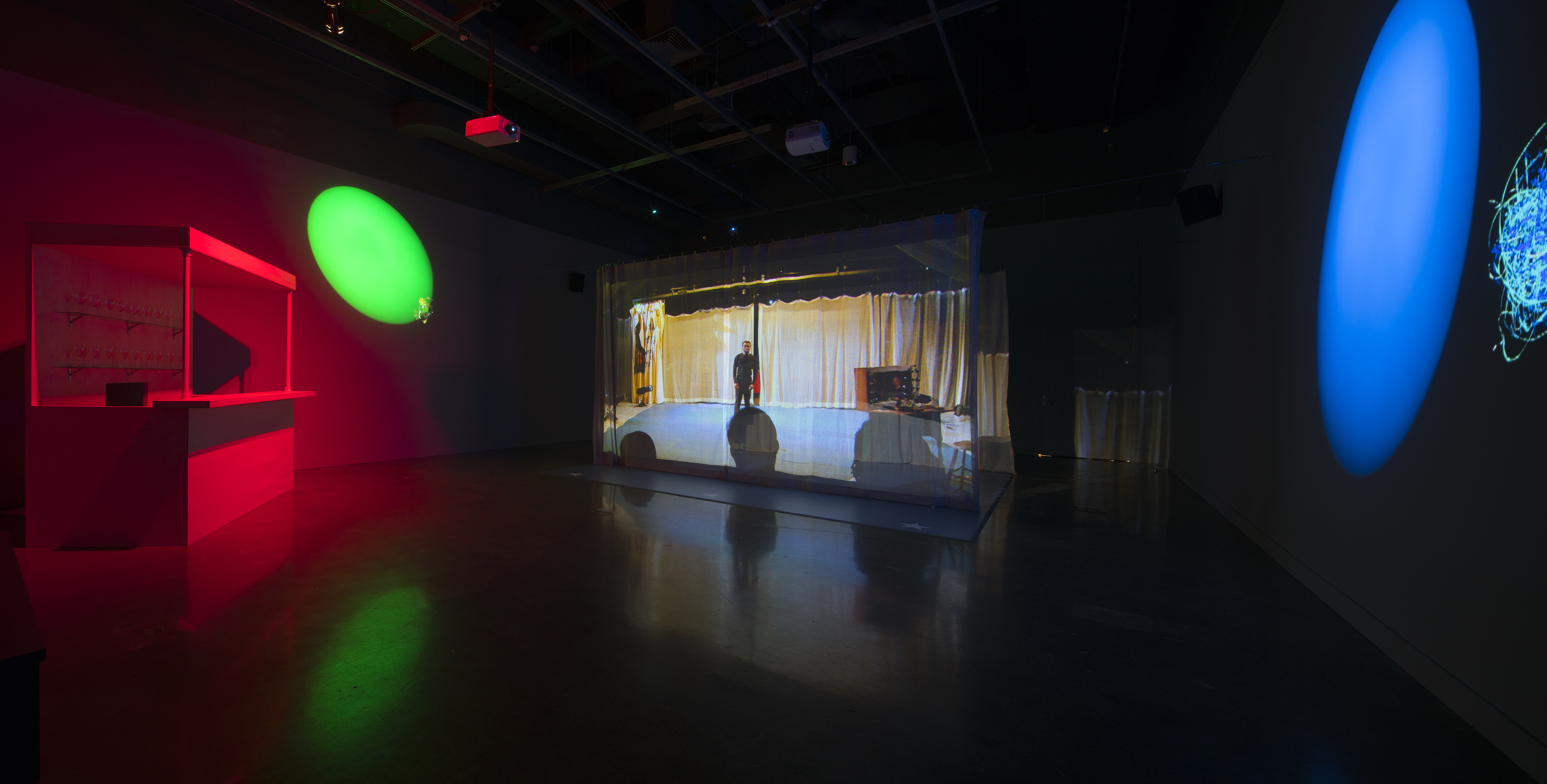 A darkened space with a video screen in the center, large blue and green oval spotlights, and a small bar bathed in red light