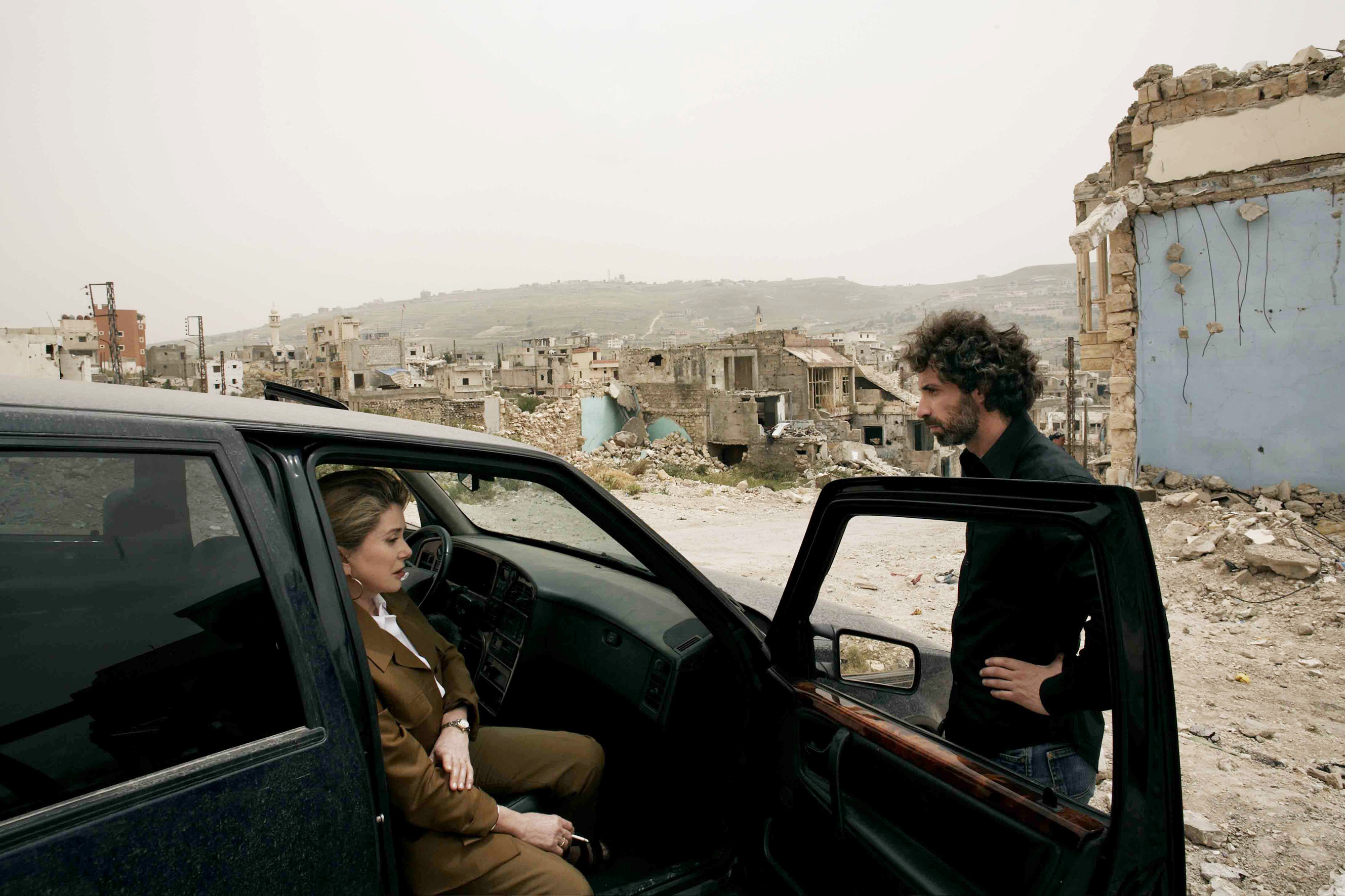 A man leans on the open passenger seat door of a black car and a woman is seated. The ruins of a city are pictured behind them.