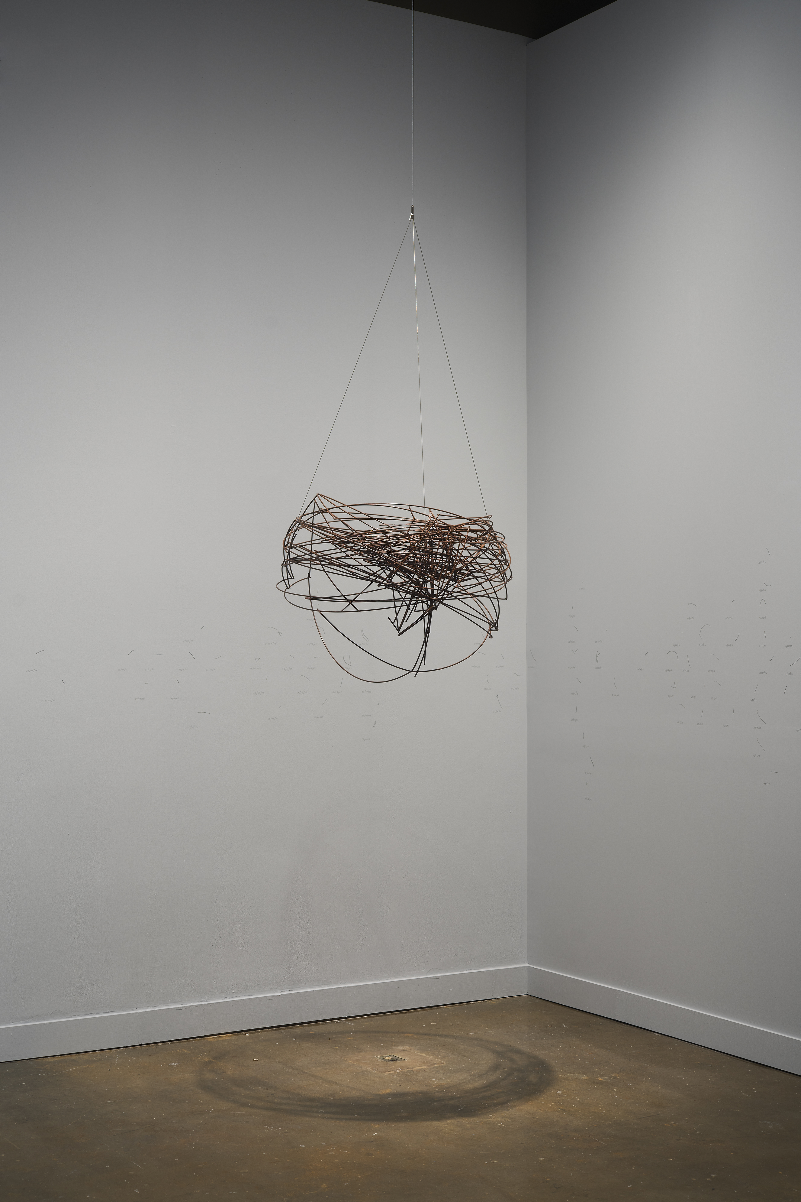 A spherical, brown-bronze-color metal-wire sculpture hangs from the ceiling before white walls in a corner of the gallery.