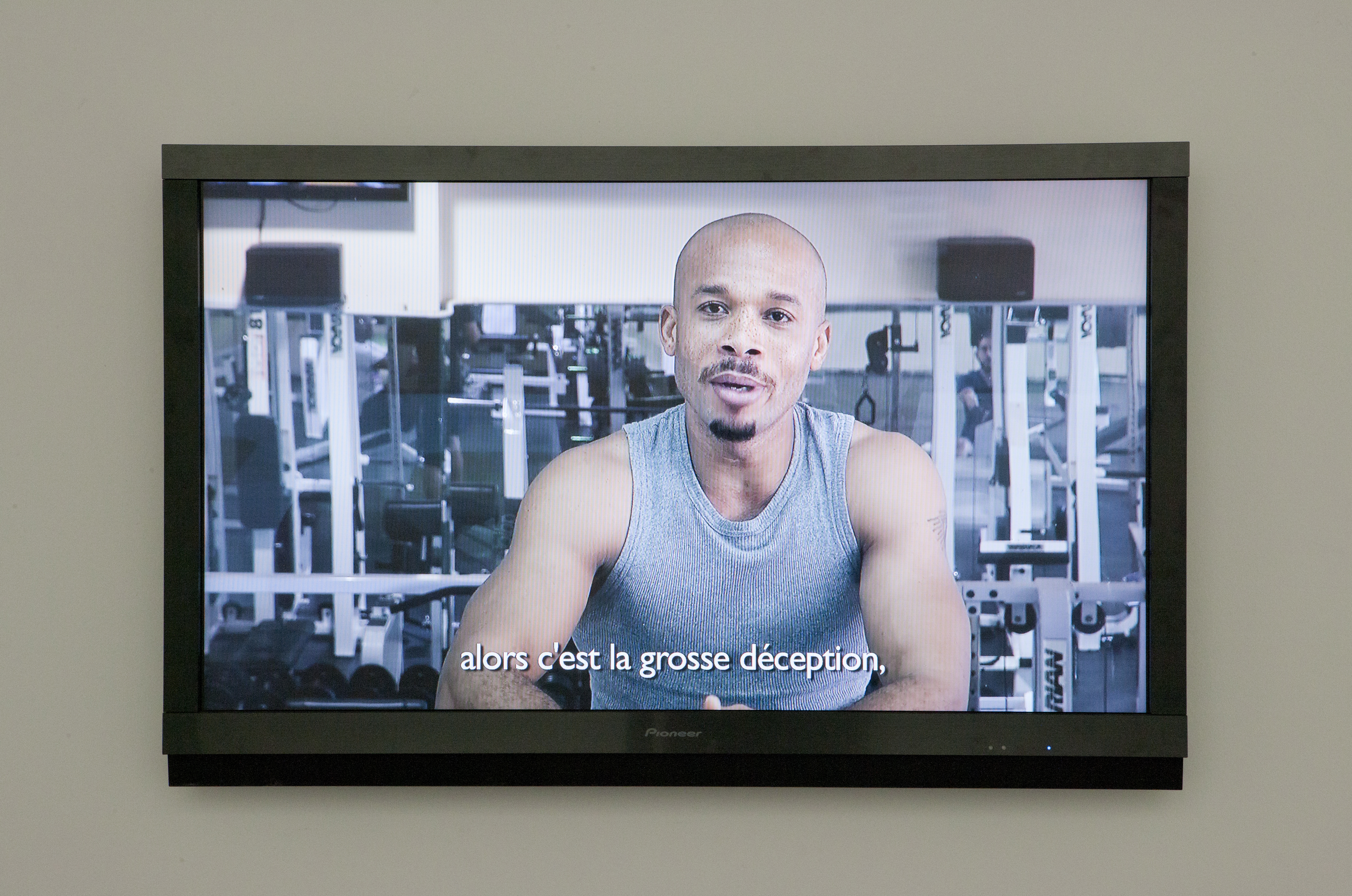Wall-mounted monitor show a muscular young man in gray tank-top in a gym with the caption “alors c’est la grosse déception,”.