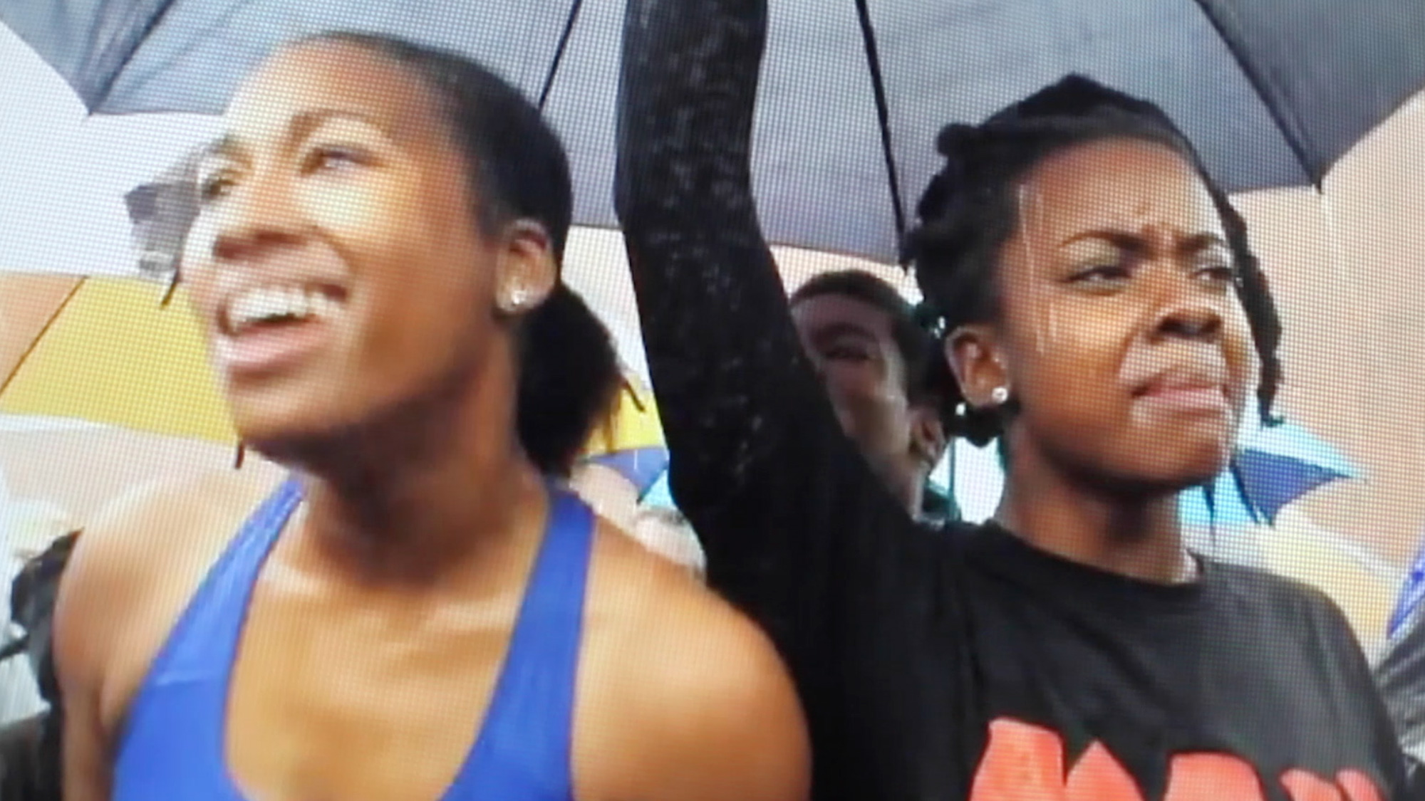 Three women stand under a black umbrella at a protest.