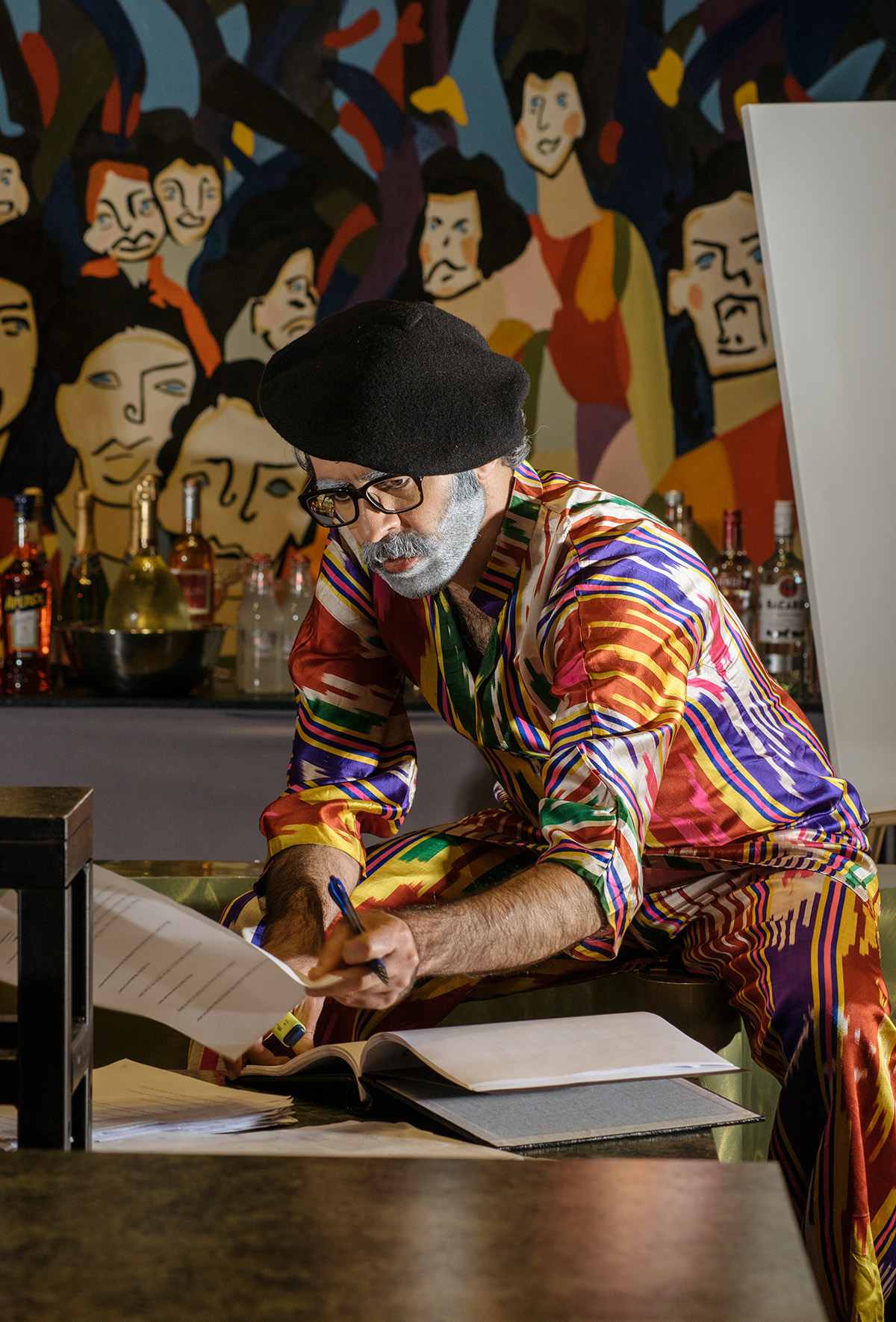 Seated actor wearing a black beret and bright striped multi-color shirt and pants flips through pages on stage.