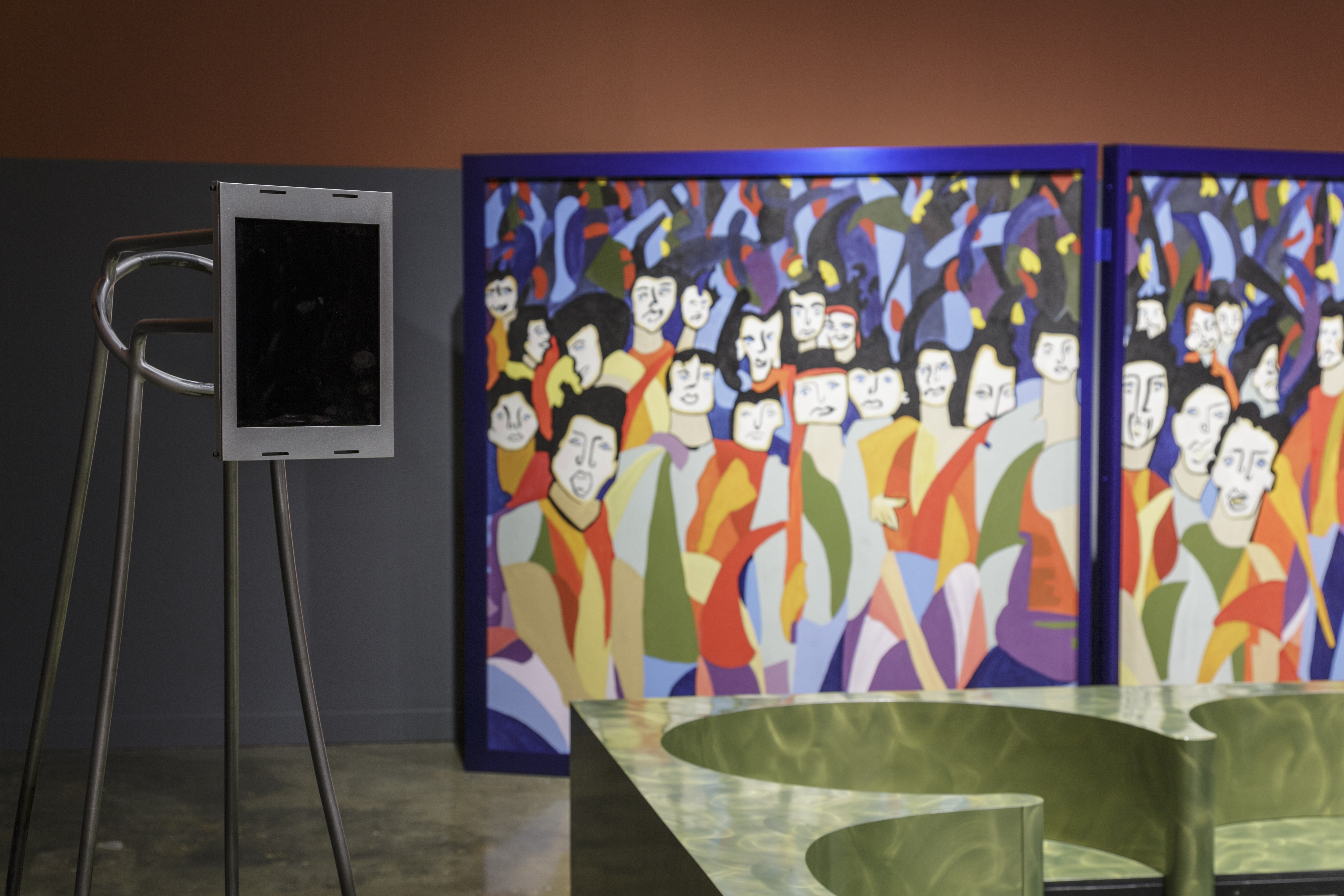 Metal upright sculpture with steel encased iPad, a corner of green banquette and colorful trifold screen in background