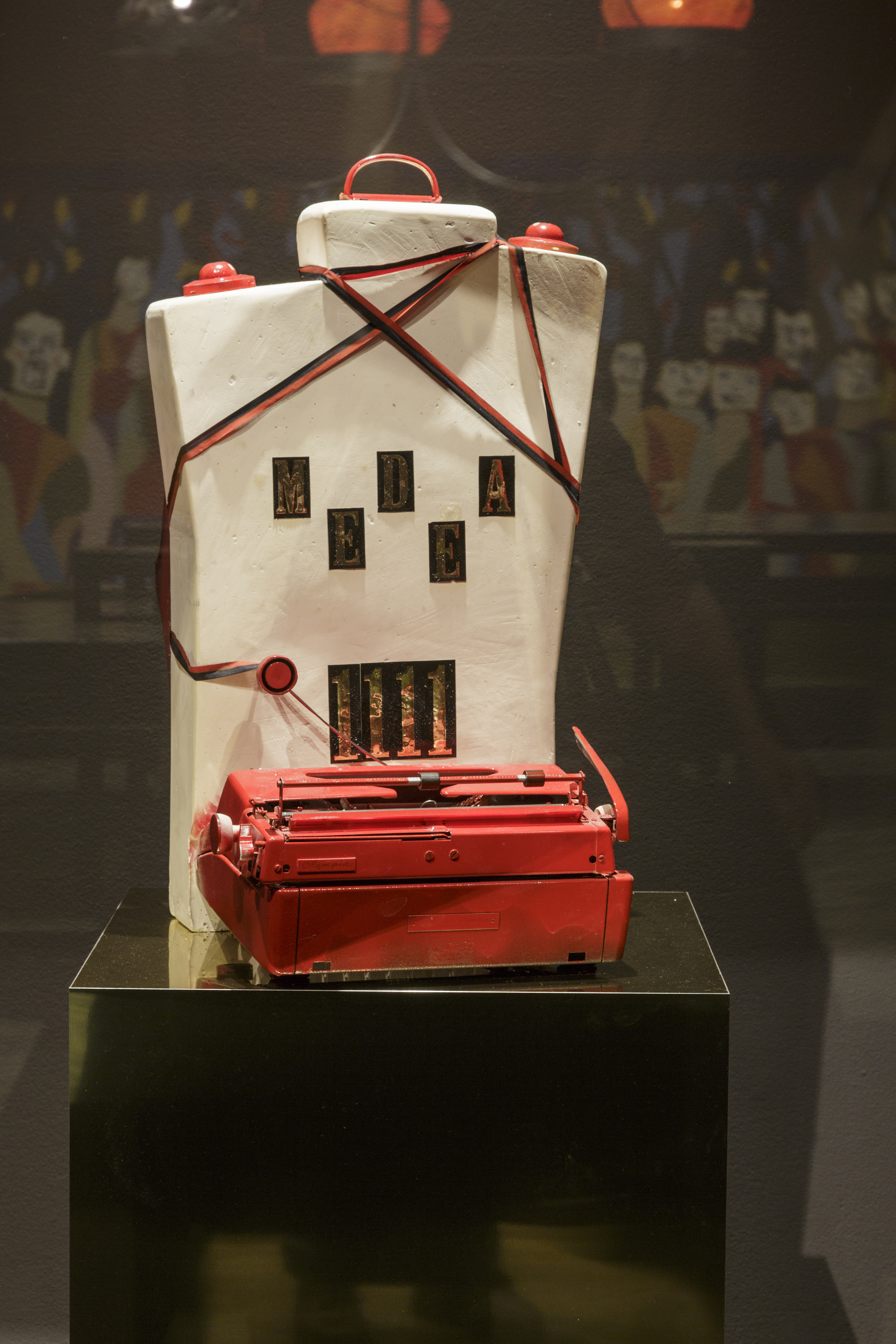 Sculpture composed of a red typewriter against a cream rectangular slab wrapped with typing ribbon on a black pedestal