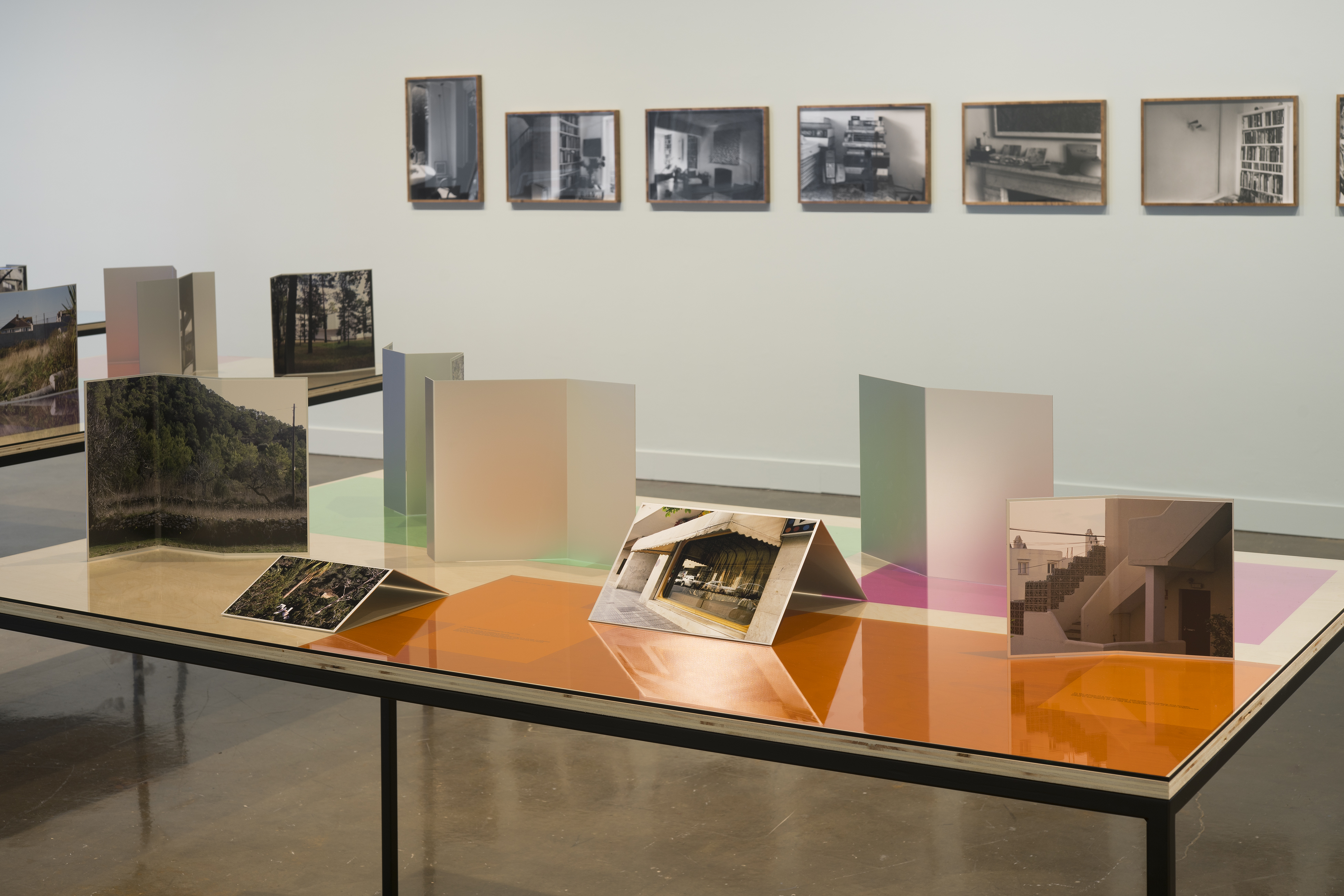 Installation view of two table tops with photographs and color gels, and a row of framed b+w photographs on the wall in back