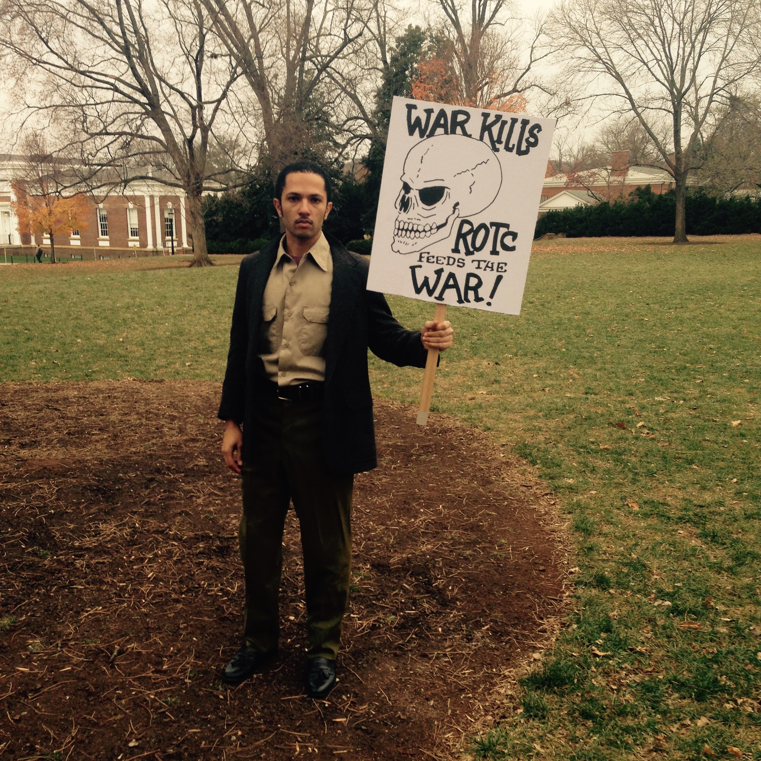 A lone man holds a sign with the words War Kills above an illustrated skull. There is a columned building in the background.