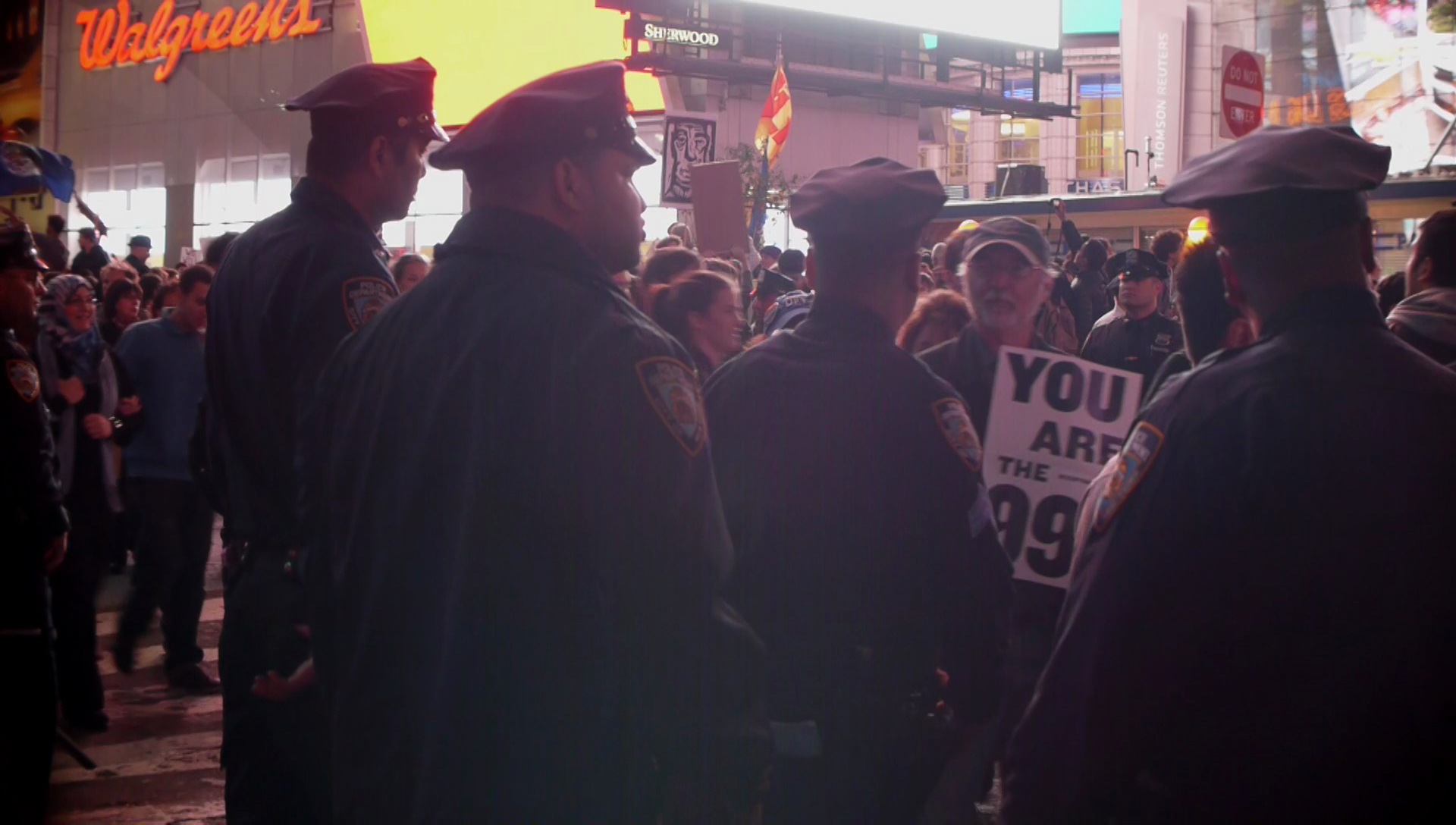 Police facing away from the camera look into a crowd on a city street. A bearded man angles a protest sign toward them.