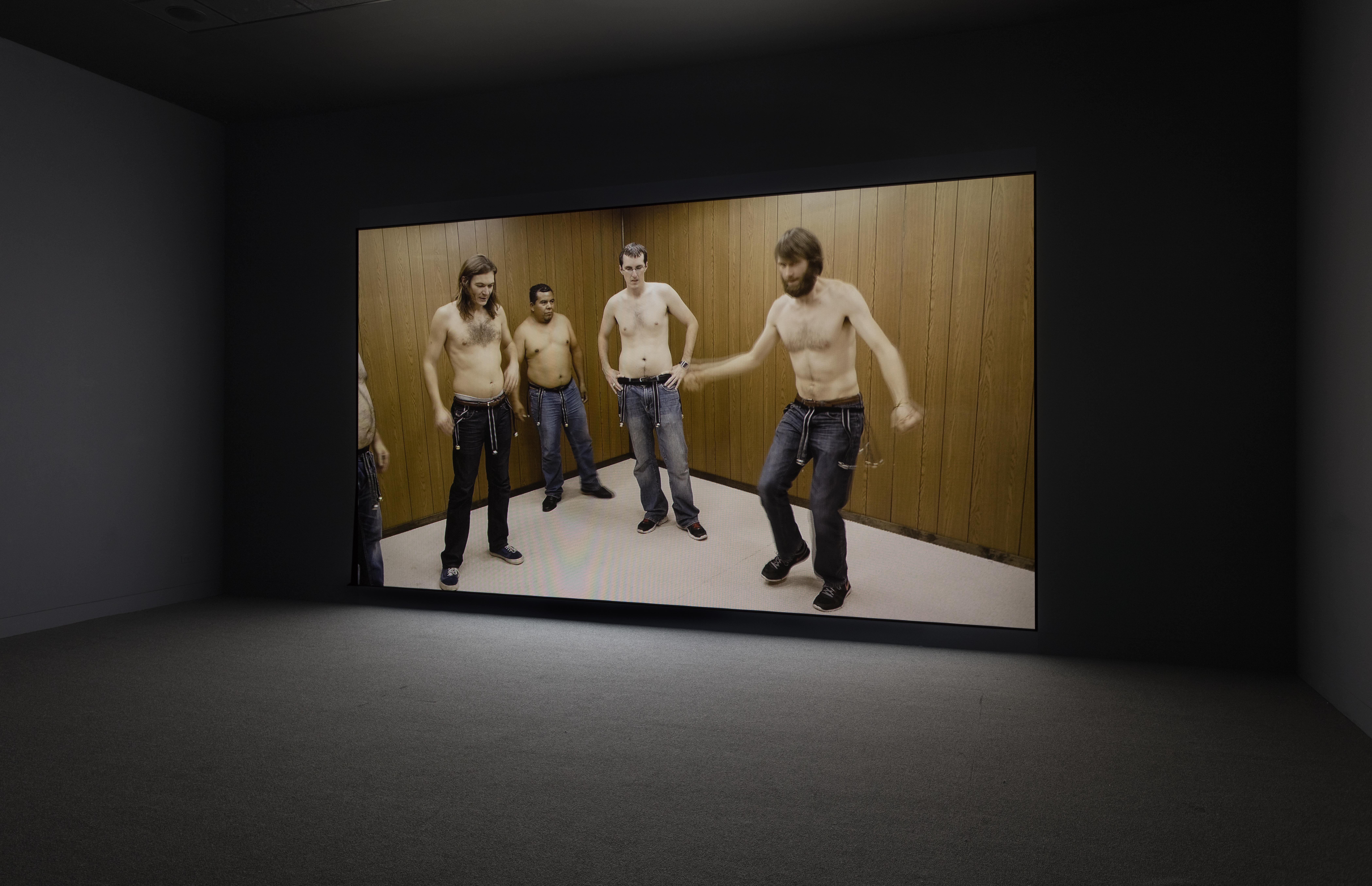 Four shirtless men, one mid-motion with arms outstretched, the other three watching, all wearing belts with bells dangling