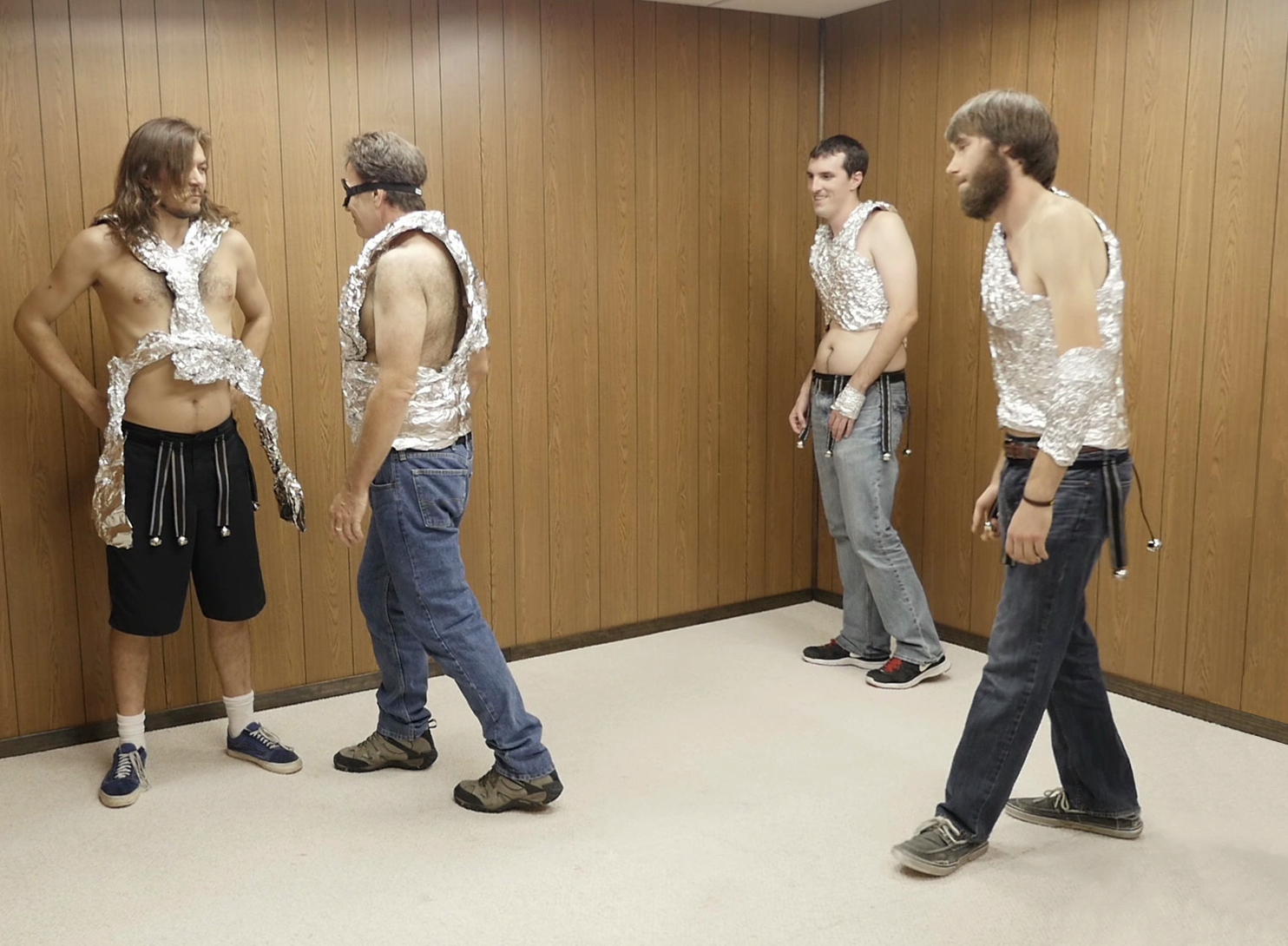 Still of a wood paneled room with four men in jeans and tin foil vests, one with his back to the wall, the others facing him