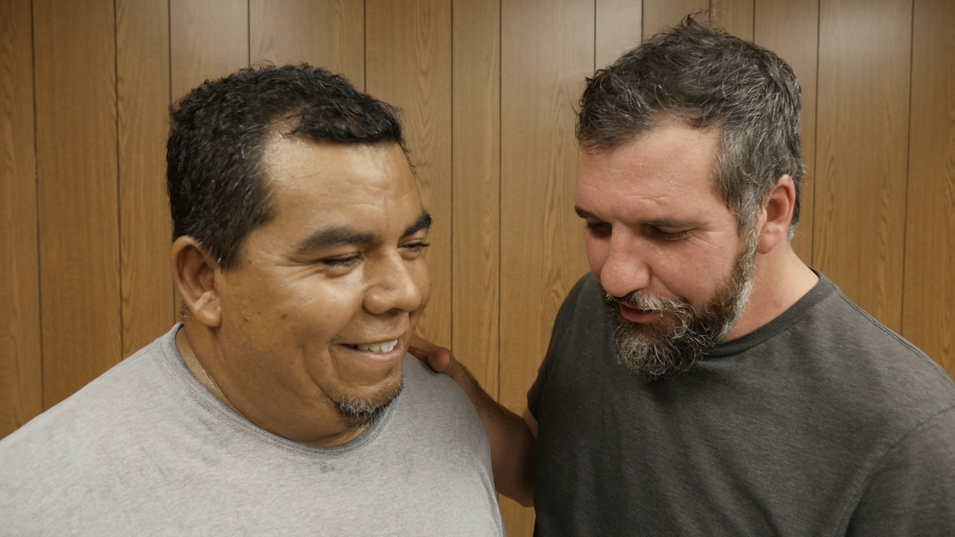 Close up of two men, one with a beard has his hand on the shoulder of a man with a goatee, who is smiling