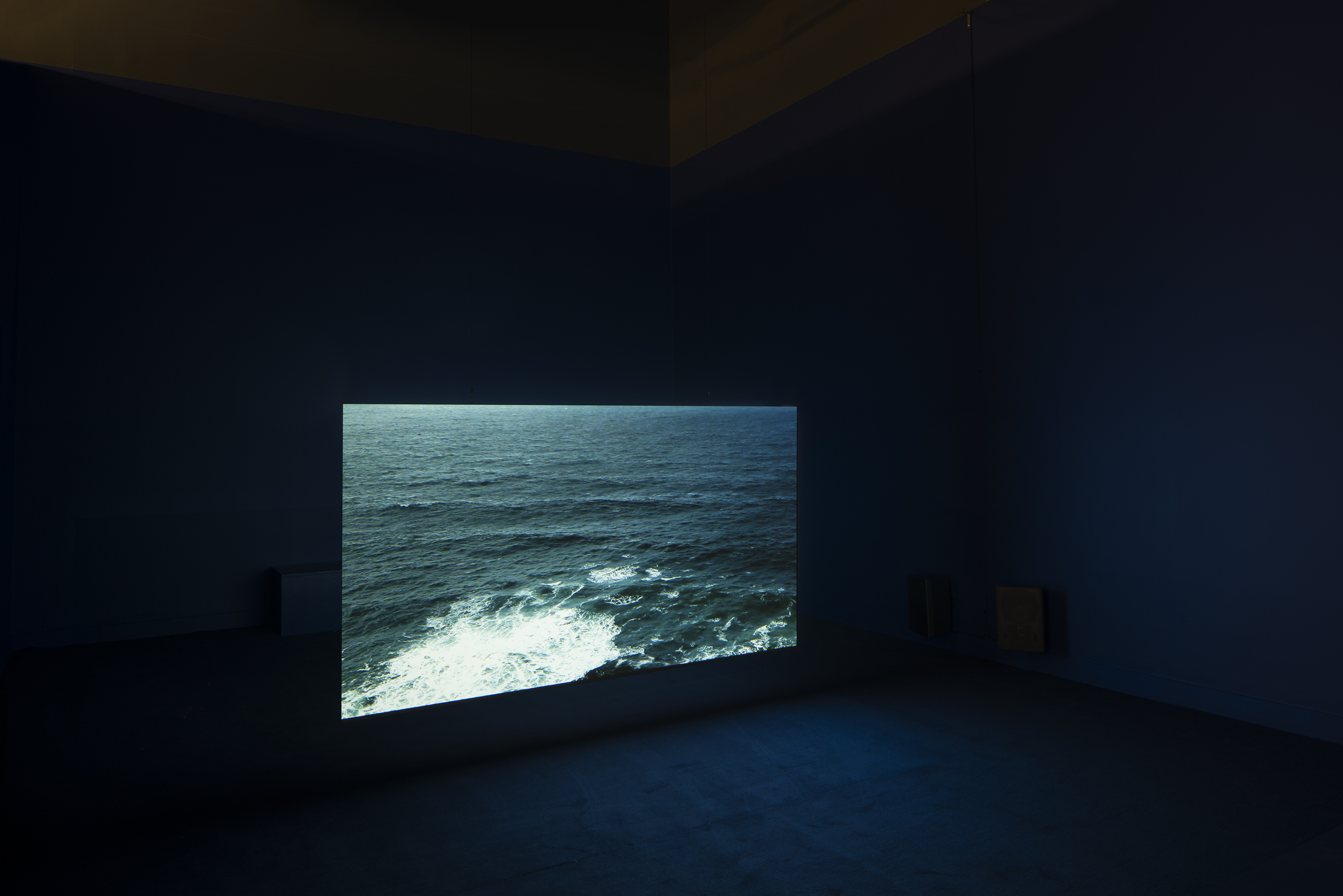 A pitch black space with a projection of a dark blue body of water with ripples and a white, crashing wave