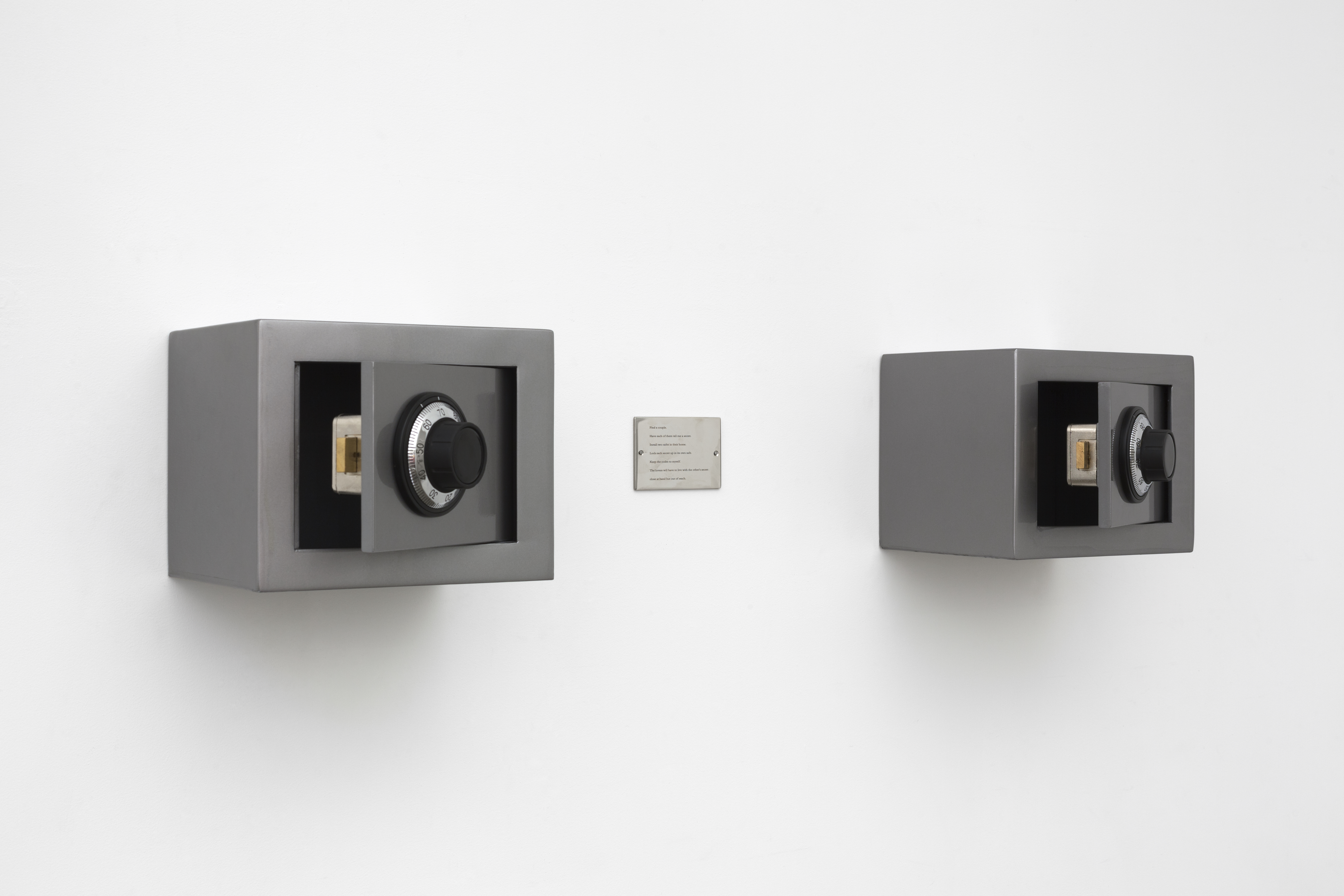 Two small rectangular safes, both partially open and secured on a white wall, an engraved plaque centered between them