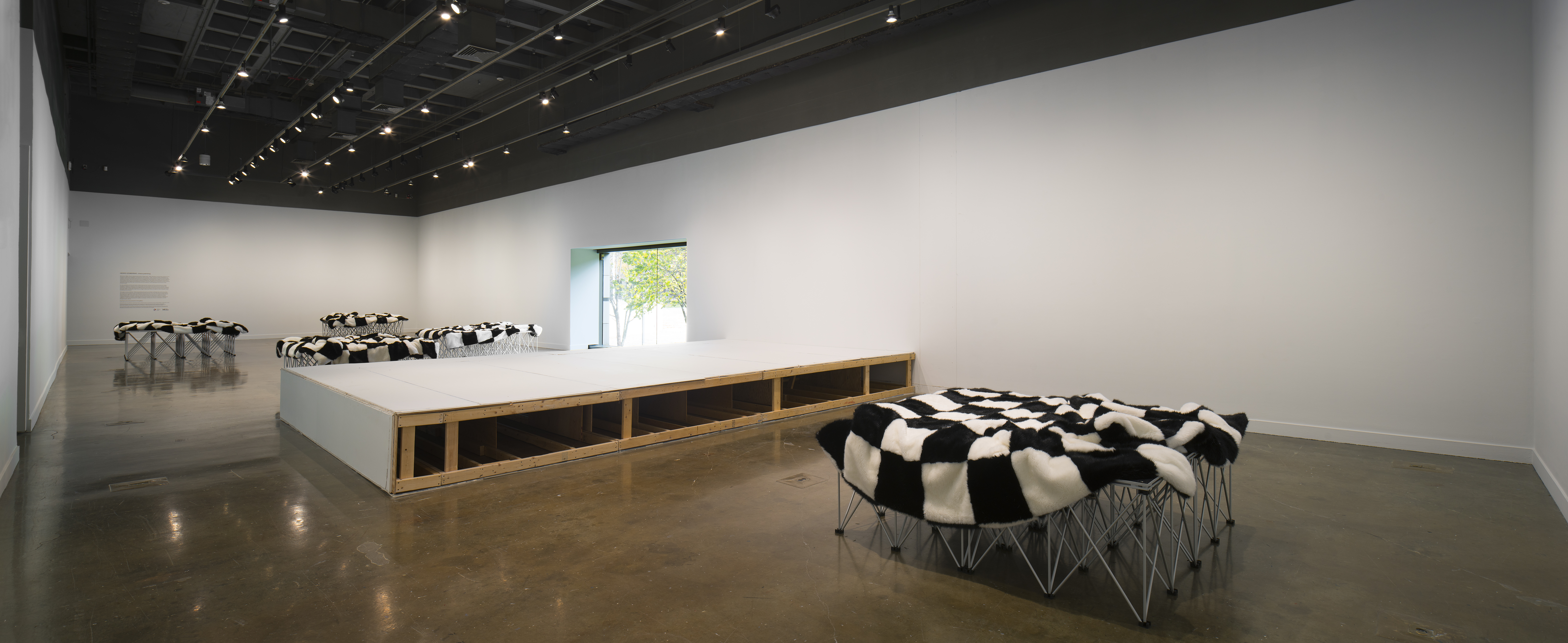 A black and white checkered fur blanket sits on multiple stilt structures in a gallery space.