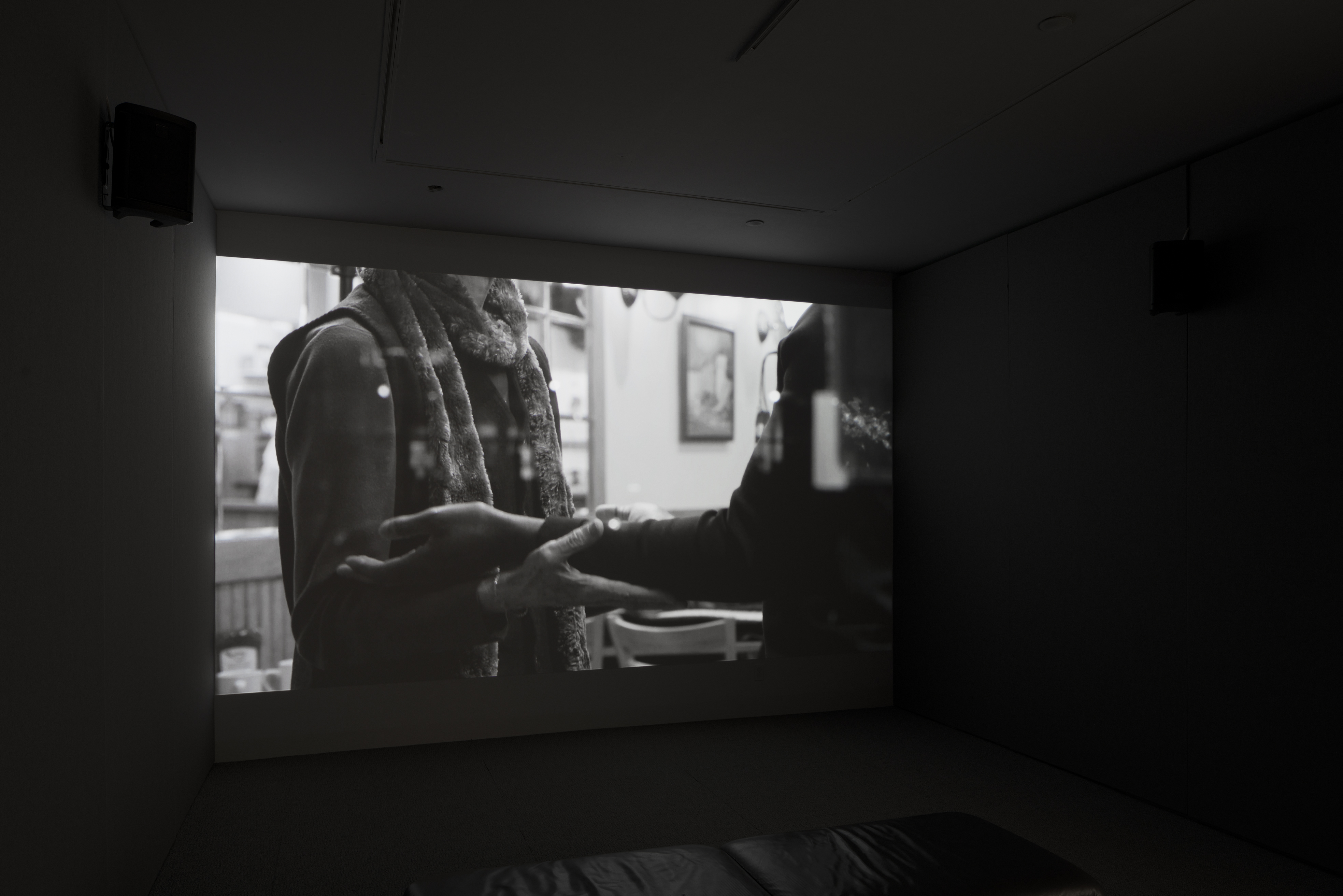 Dark room with video projection of a person's torso, arms bent at right angle, palms up, supporting the arms of another.
