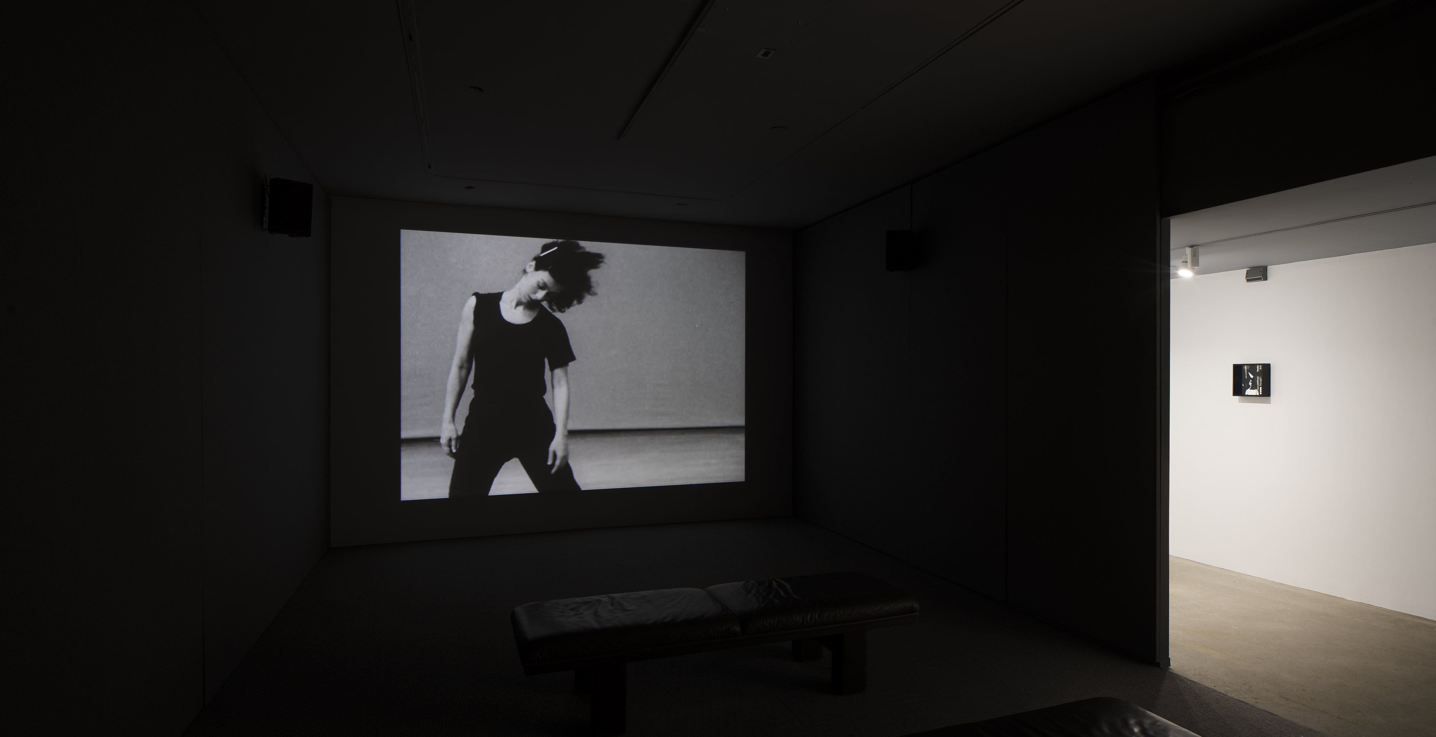 A dancer in motion is projected in a dark room. An opening leads to a bright hall where one small work hangs.