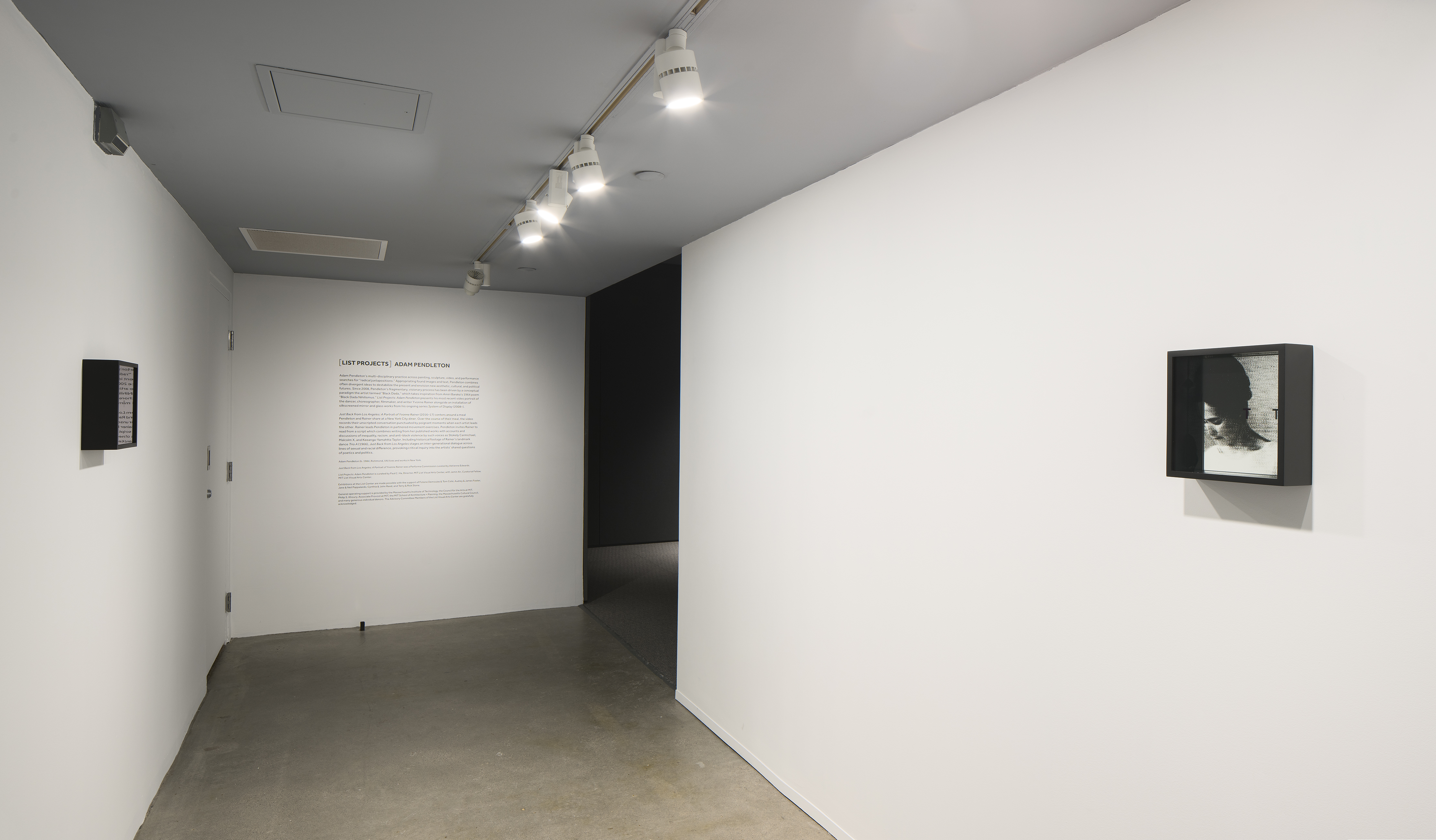 Corridor with track lights, small works on left and right walls. Wall of text at the far end leads into a darkened room.