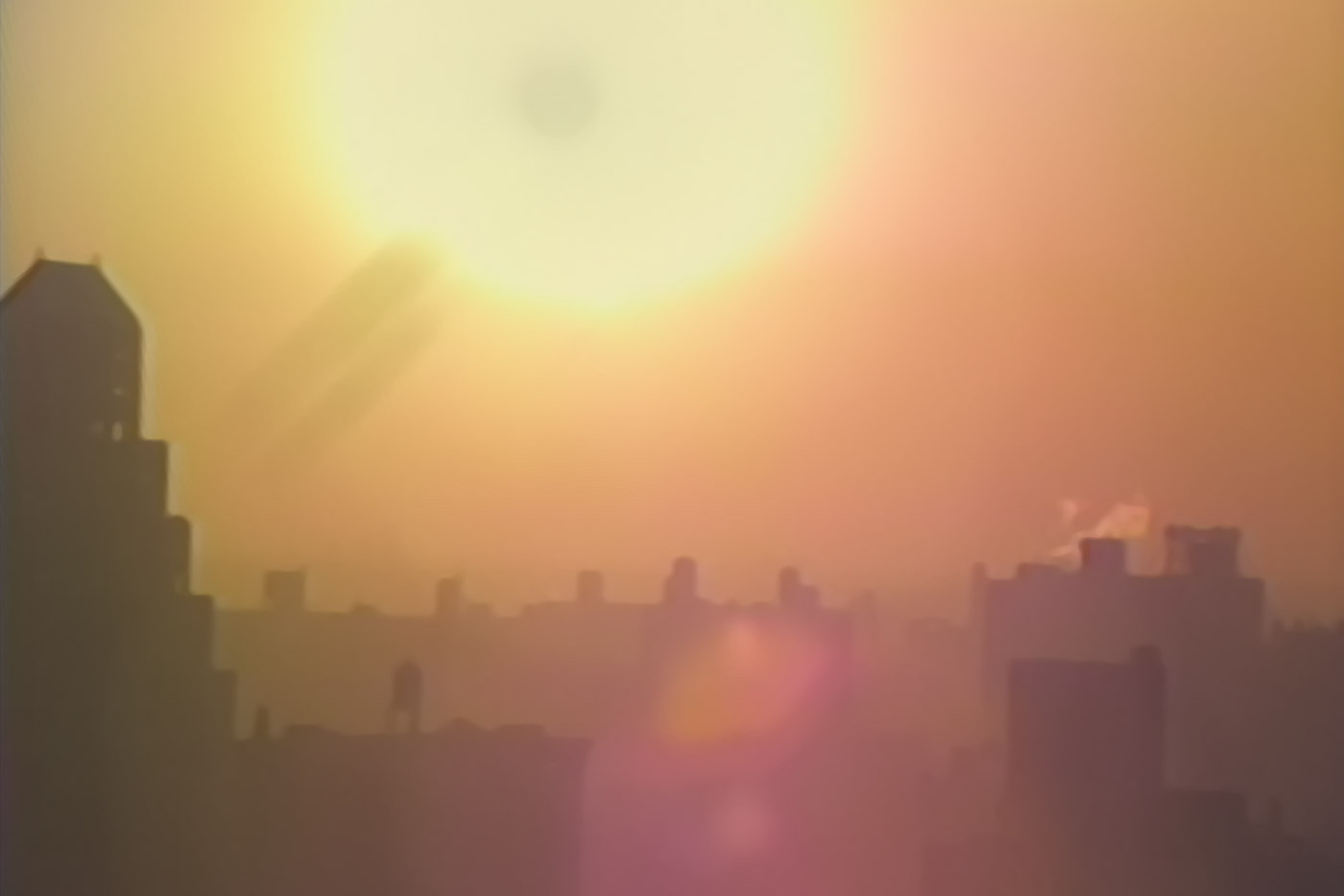 Still of a blurred sunrise, casting orange and pink, fog like, over city rooftops.