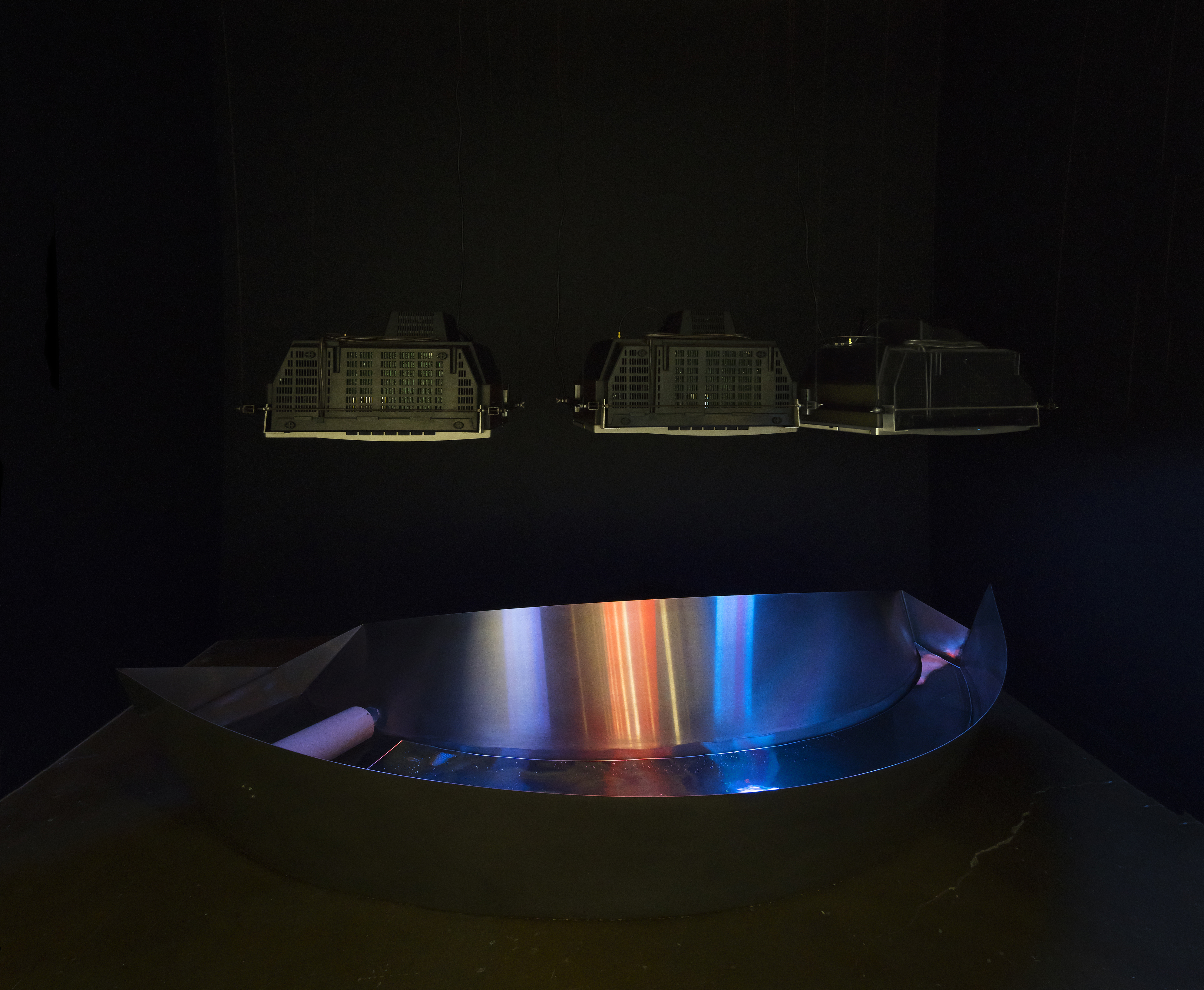 A curved, steel trough reflects colored images from 3 CRT monitors, suspended face-down above it, in a dark space.