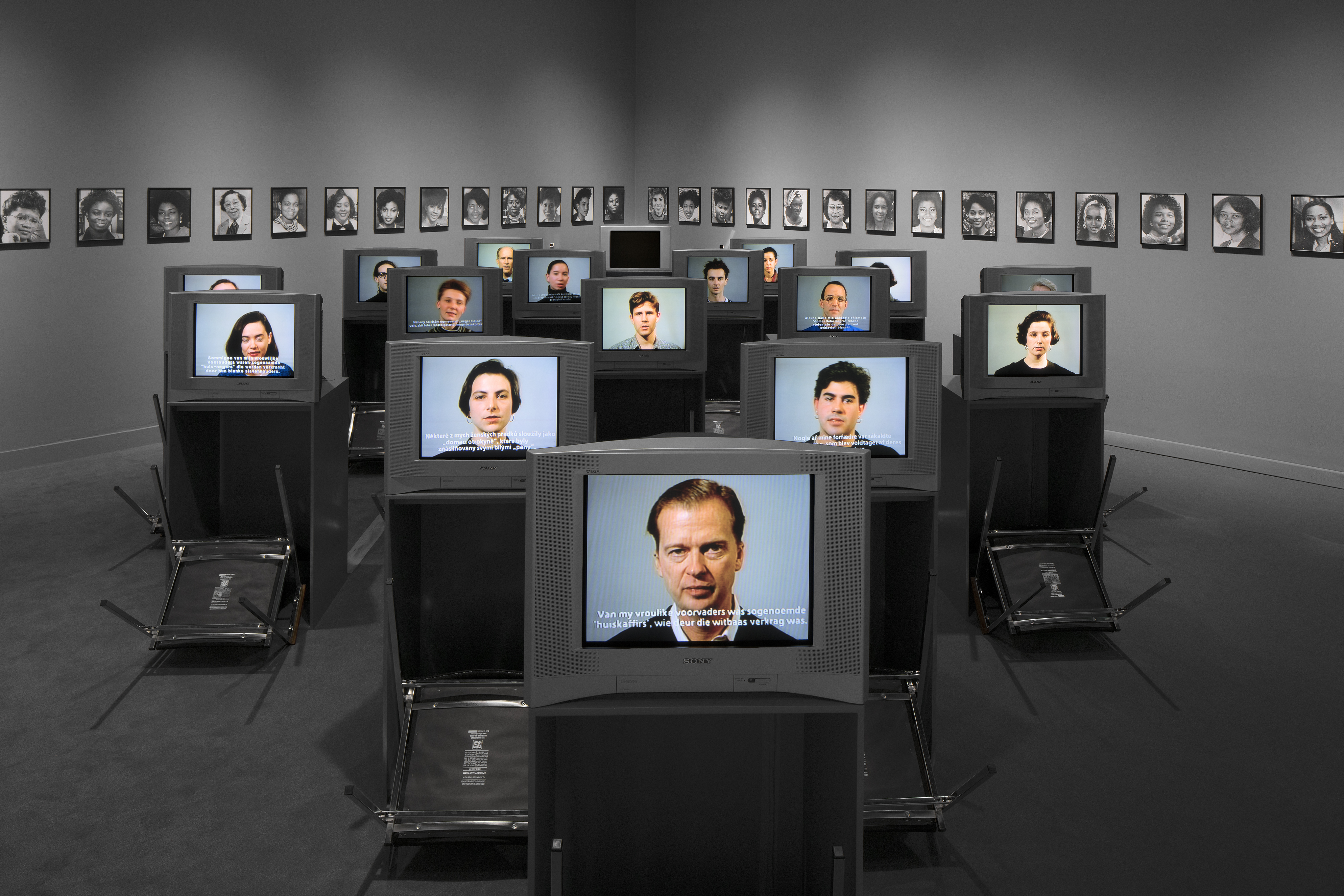 An array of video monitors on desks displaying faces, upside down chairs placed before each, portraits line the walls in back.