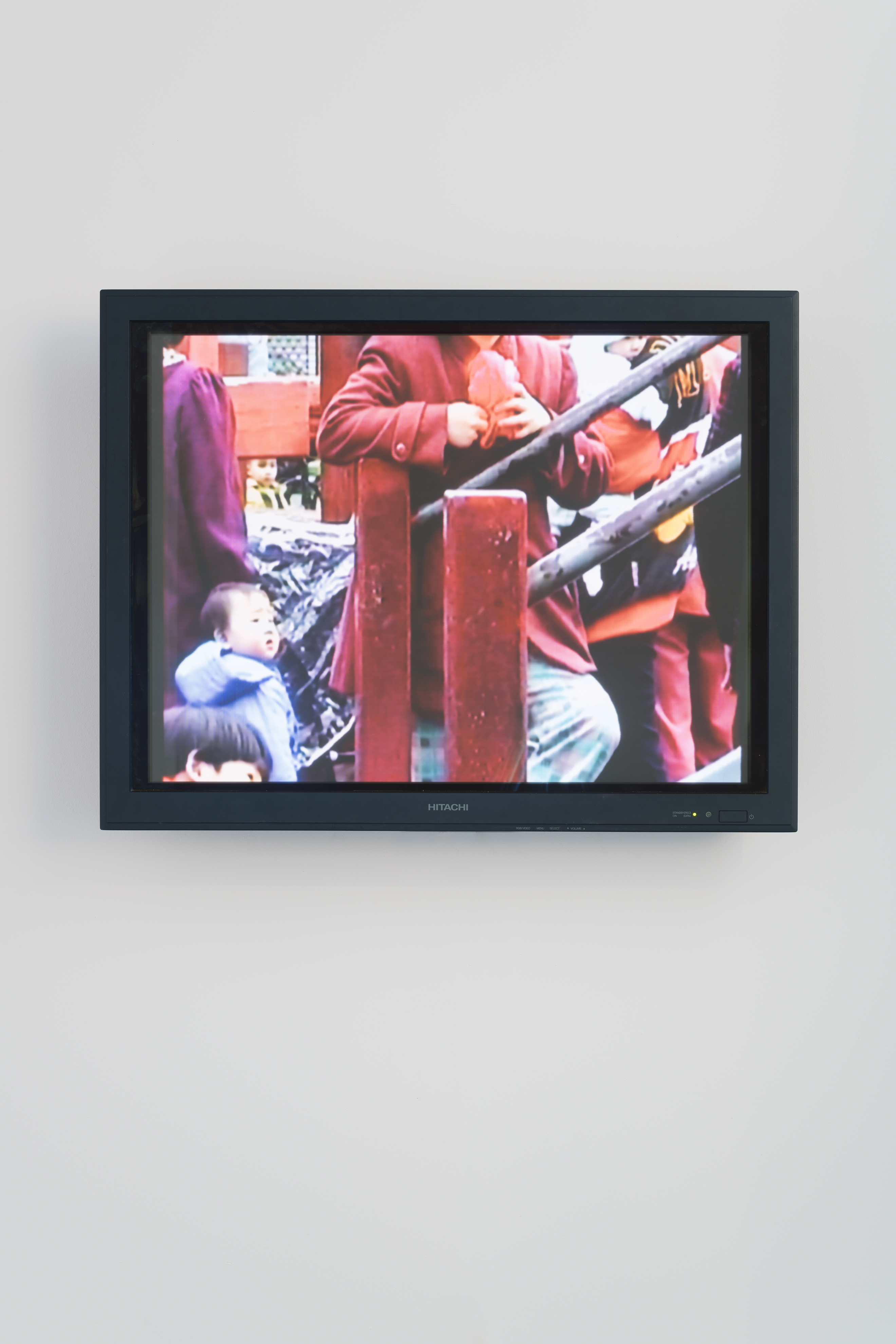 Video monitor shows a colorful scene of a torso in a red coat behind a railing, a small child, glimpses of others around them.