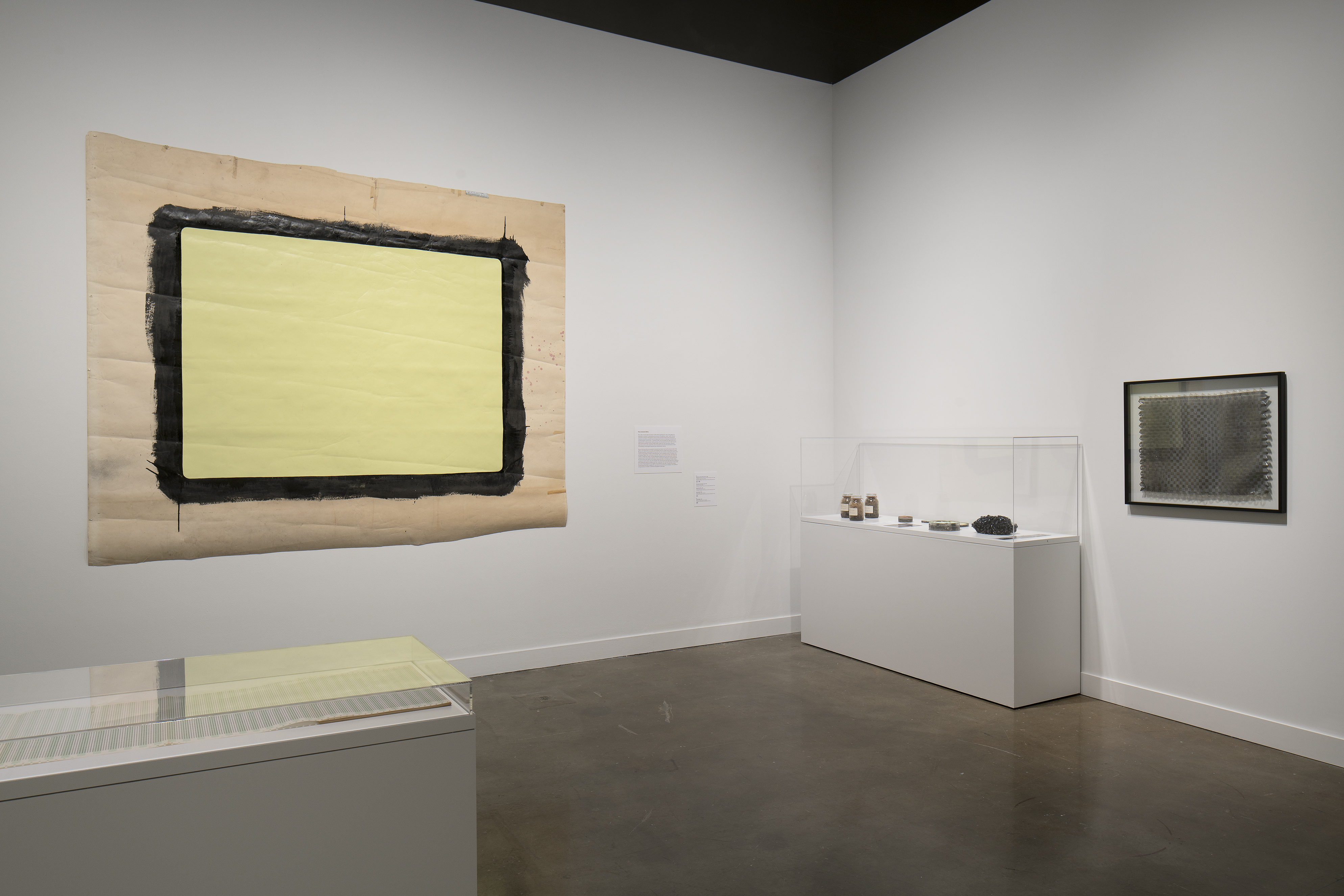 Two glass-topped vitrines are flanked by a large mounted painted piece of paper and a framed object on perpendicular walls.