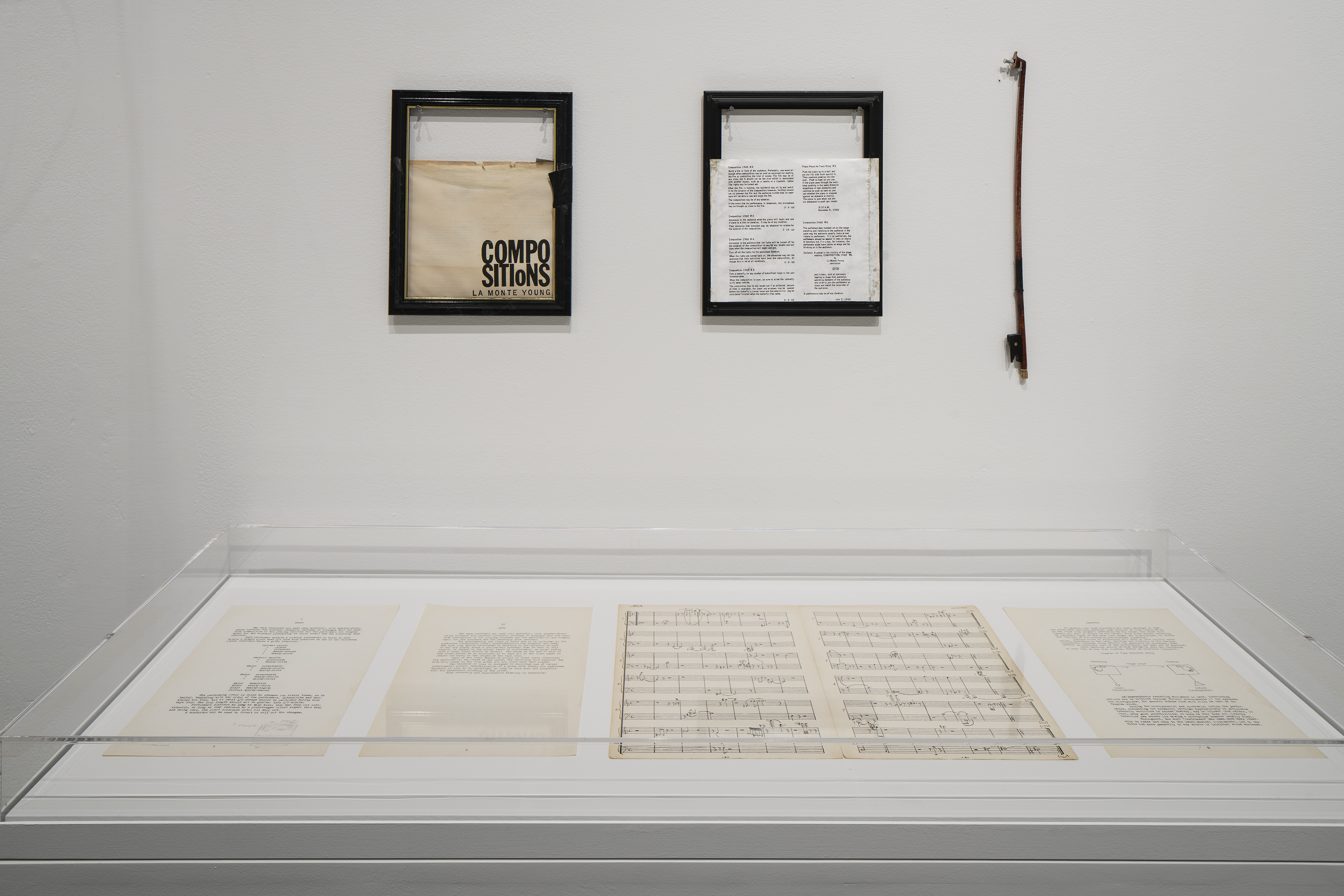 Two framed objects and a half-sized bow hang on a wall behind a glass vitrine containing a musical score and other printed pages.