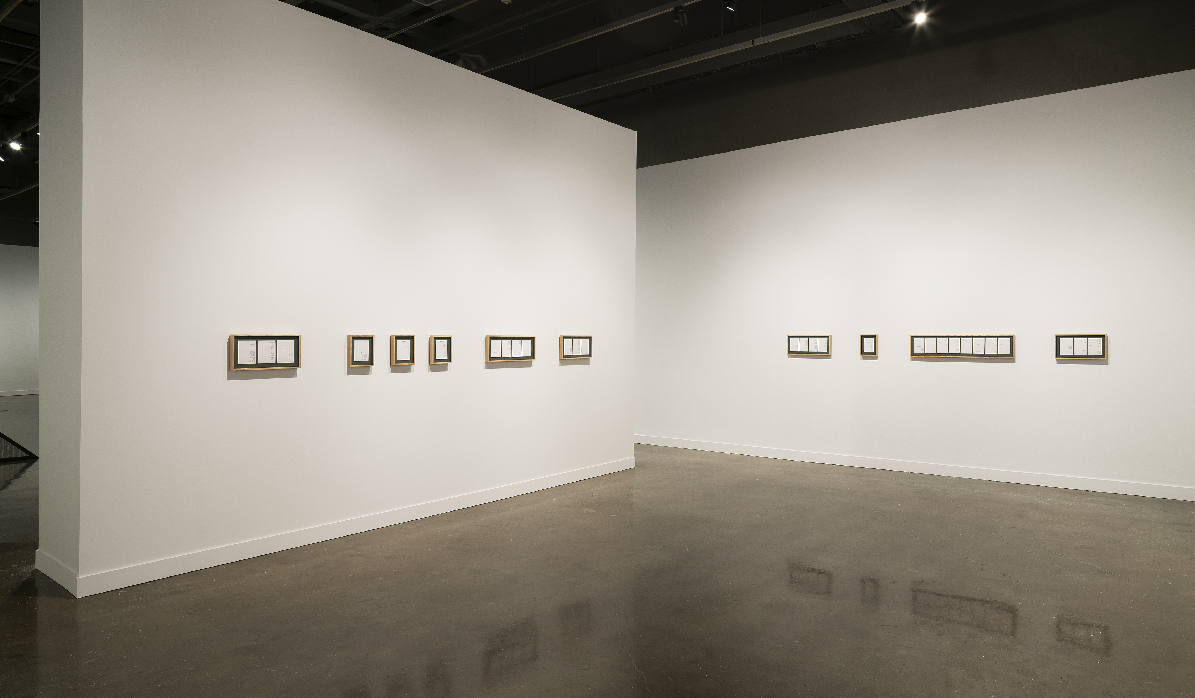 Nine horizontal frames with dark matting and white pieces of paper hang from two nearly bisecting white walls.