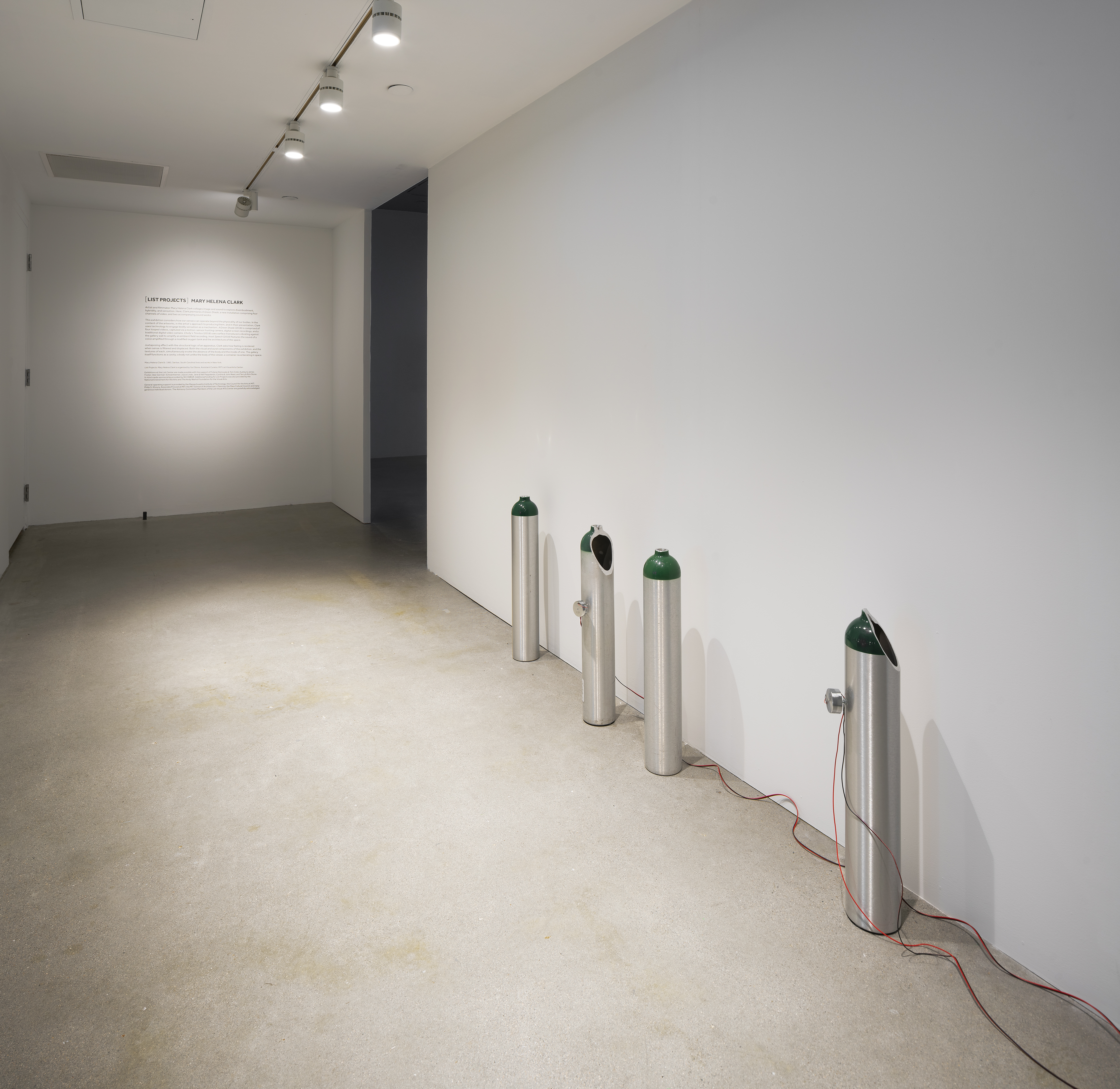 On the right side of a hallway sit 4 cylindrical tanks with painted green tops, some with holes and wires. Text on back wall.