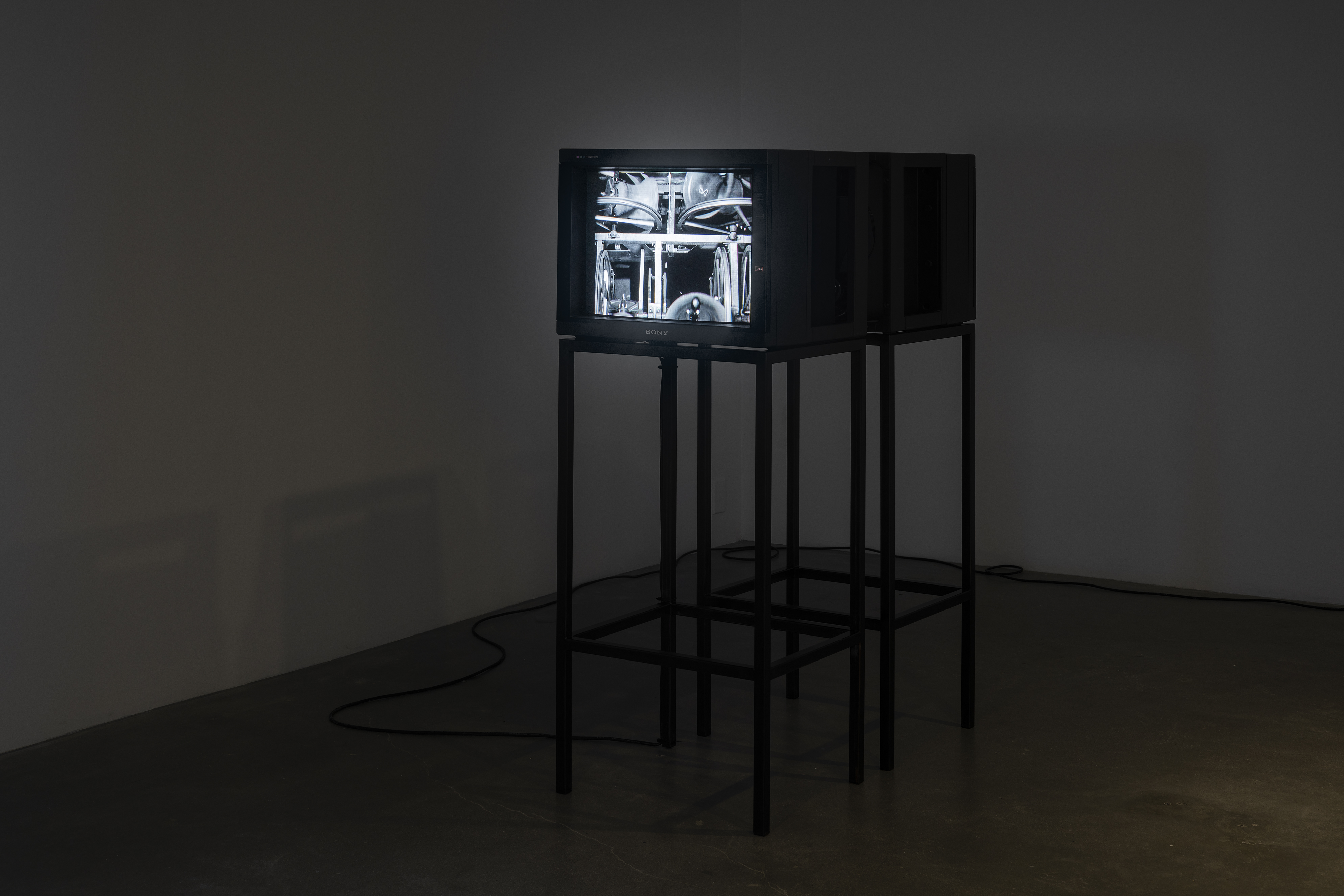 Two video monitors on stands face in opposite directions in a darkened room. A black and white image is visible on one monitor.